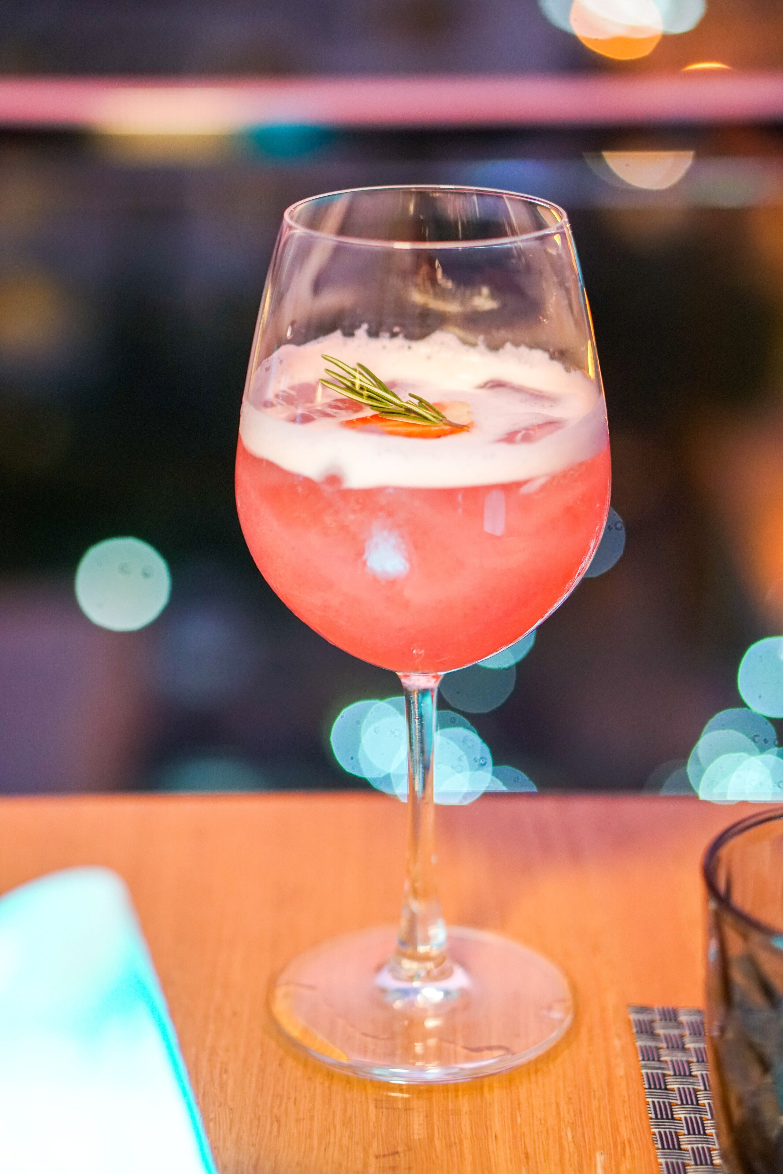 Un verre à cocktail contenant une boisson à plusieurs couches, décoré d’une branche de romarin, posé sur une surface en bois.