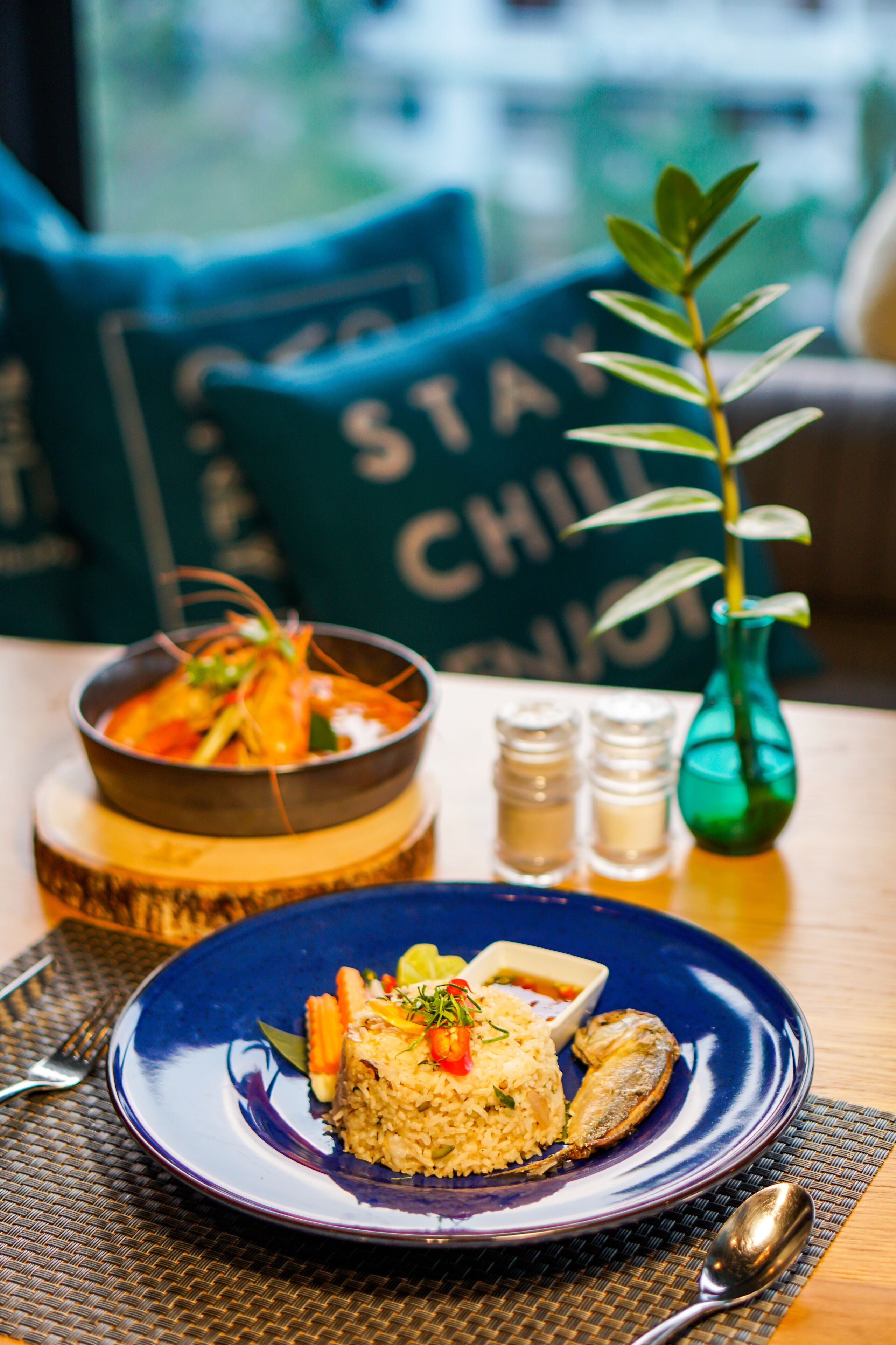 A dining table with a blue plate of food, a bowl of soup, and a green plant in a vase.