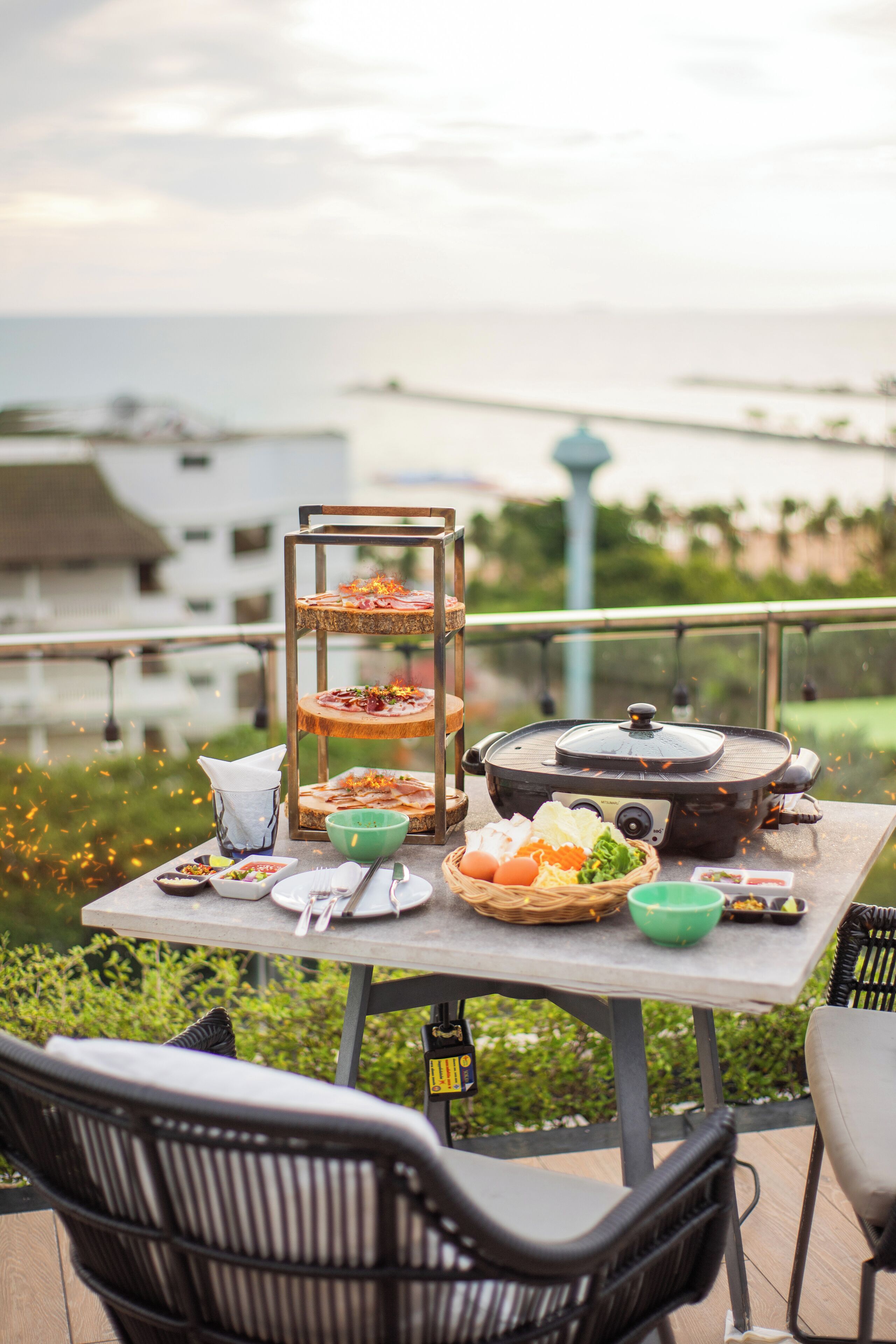 Una mesa de comedor con una variedad de platos, un soporte de tres niveles y una estufa portátil en una terraza al aire libre con vistas al mar.