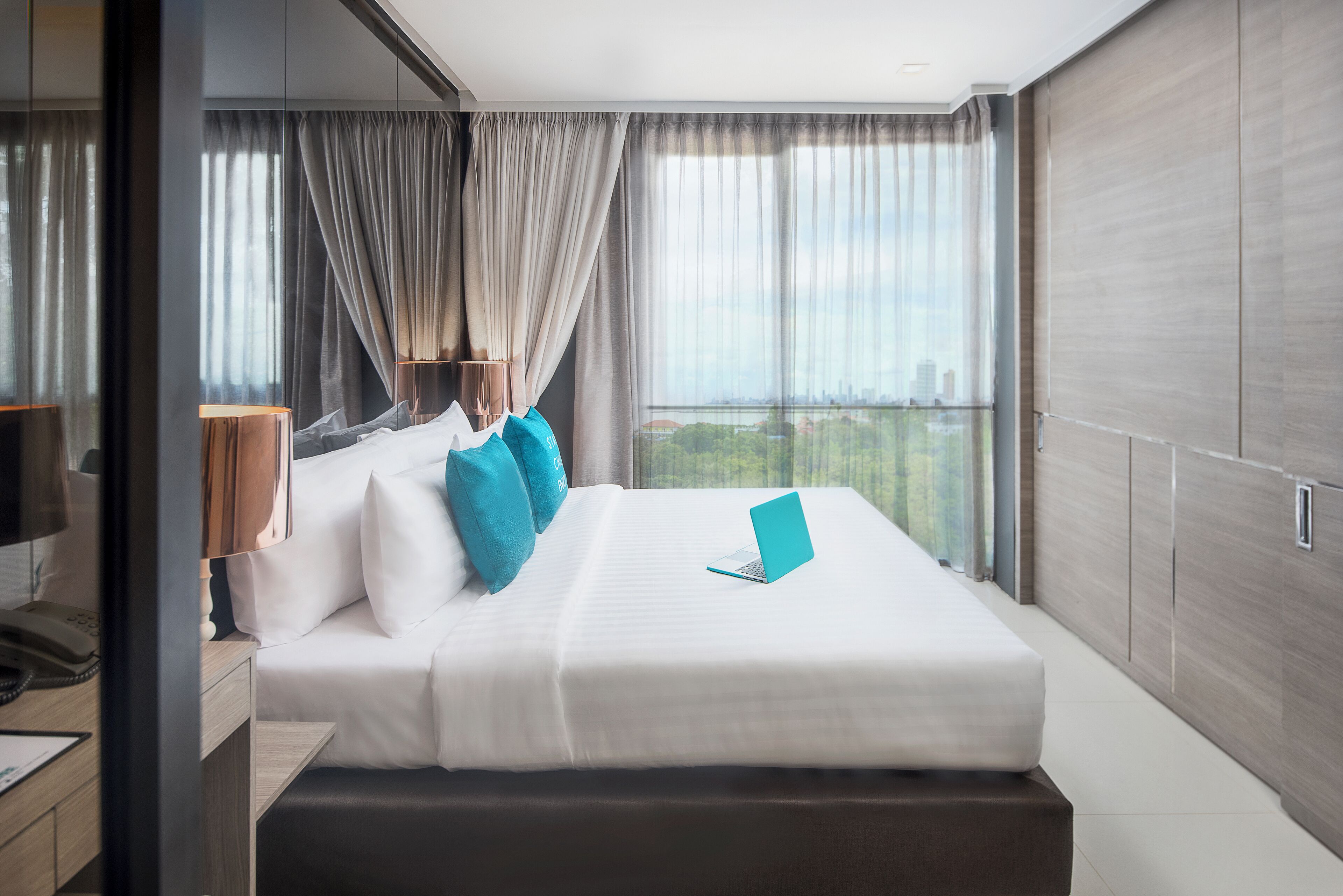 A modern hotel room with a large bed, a laptop on the bed, and a city view through the window.