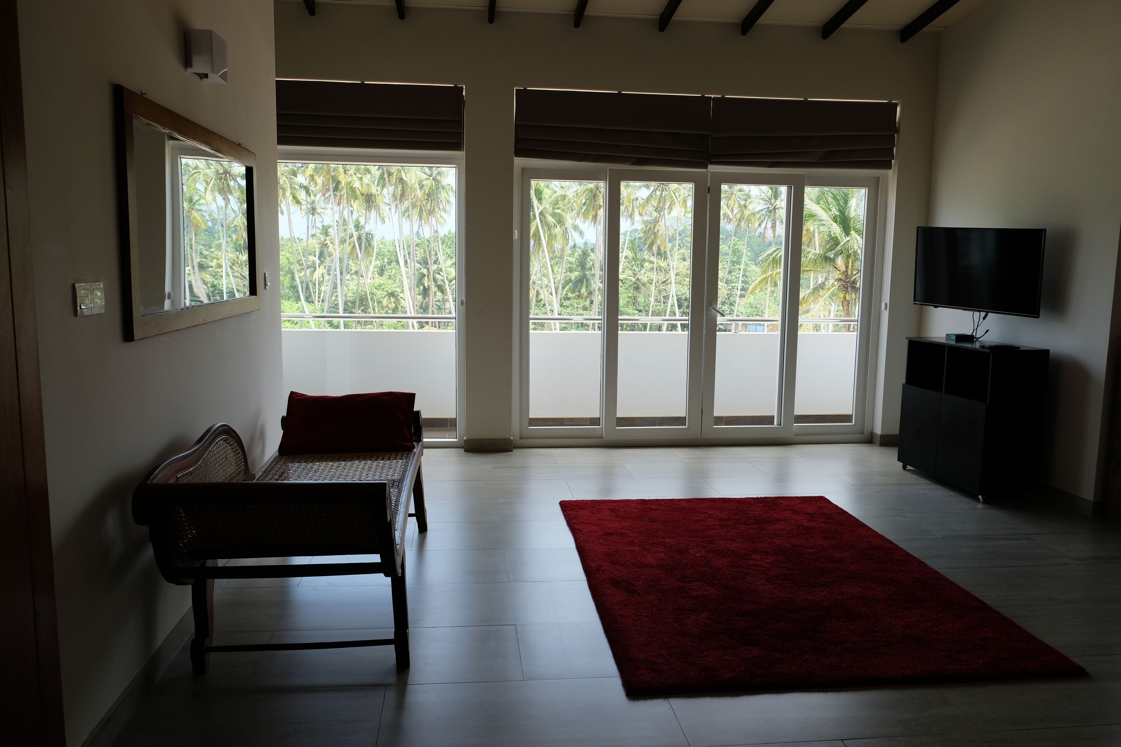 A room with a bench, a red rug, a TV on a stand, and large windows with a view of palm trees.