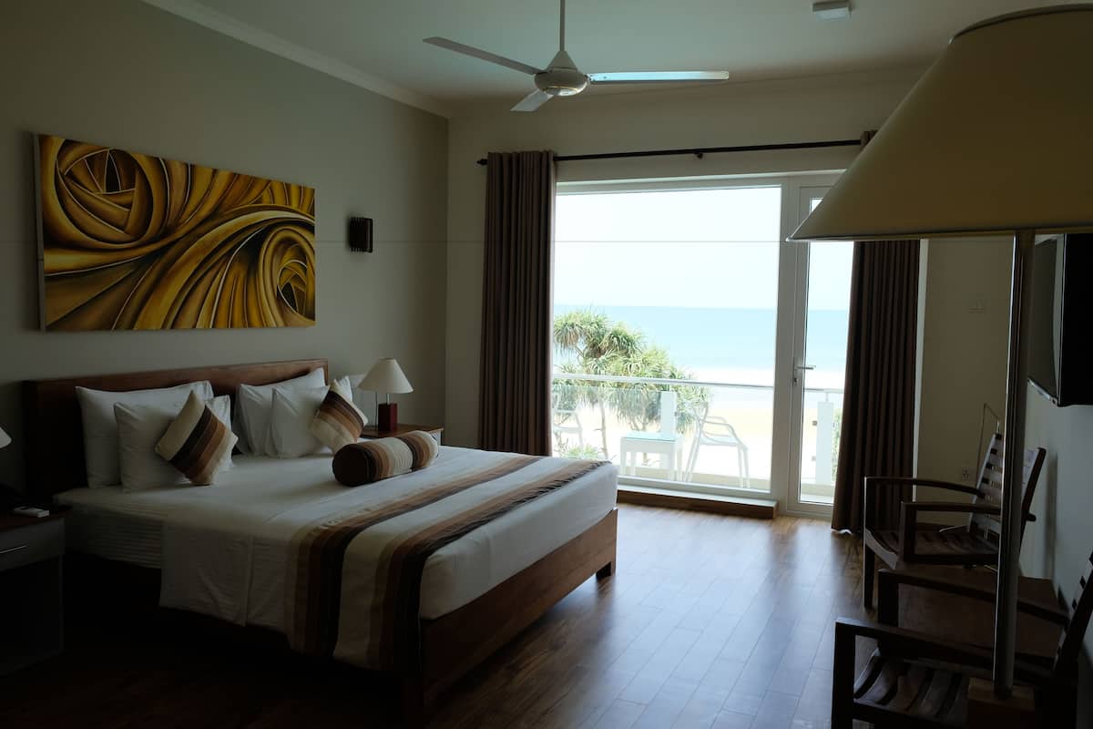 A hotel room with a bed, a ceiling fan, a large window with curtains, and a view of the ocean.