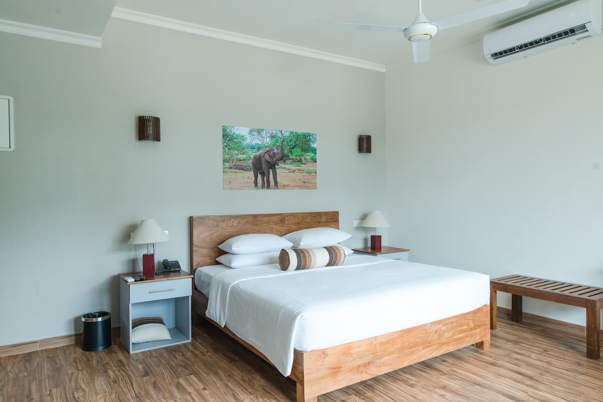 A bedroom with a wooden bed, white bedding, a nightstand with a lamp, a small wooden bench, and a picture of an elephant on the wall.