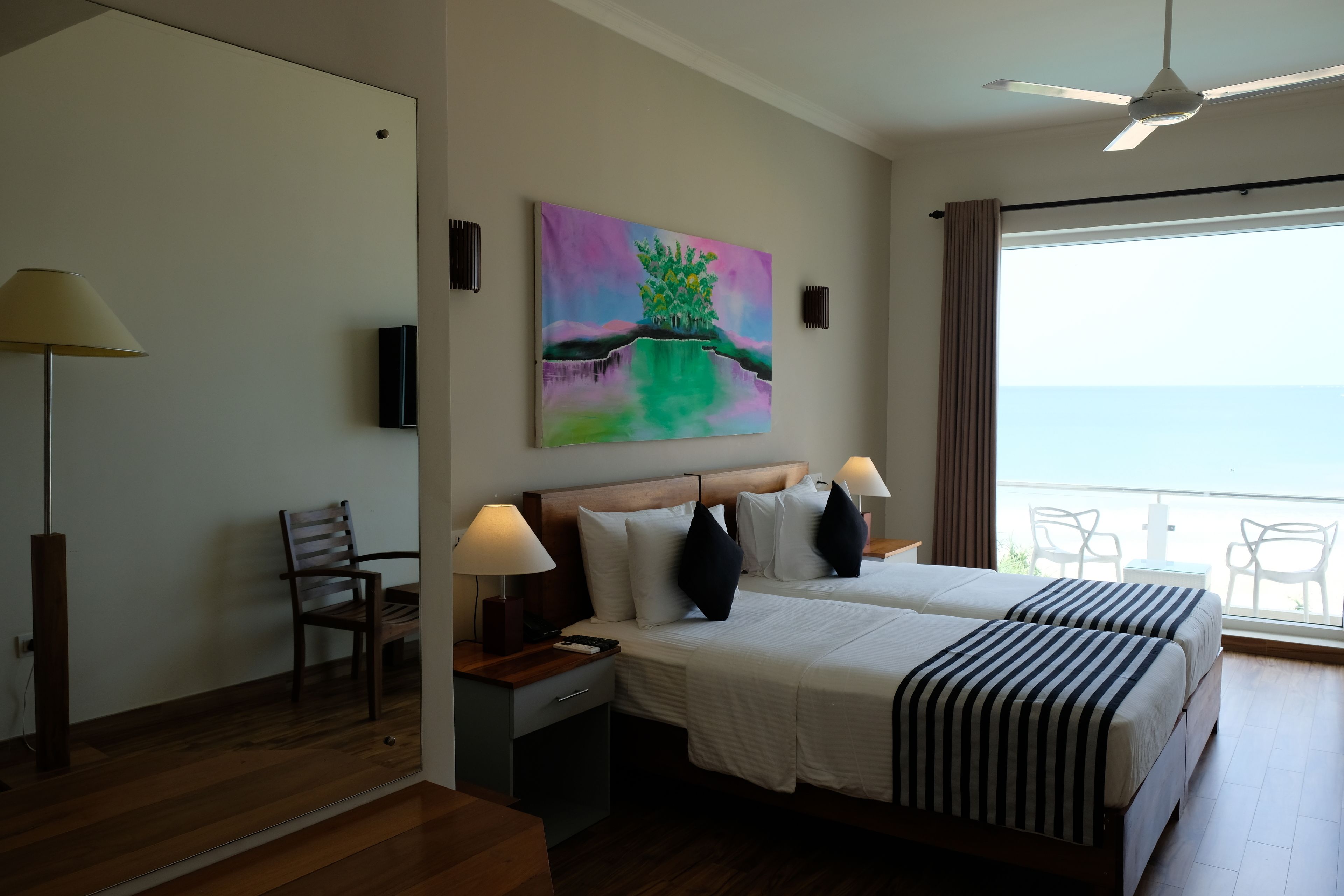 A hotel room with a large bed, two bedside tables with lamps, a chair, a painting on the wall, and a balcony with chairs.