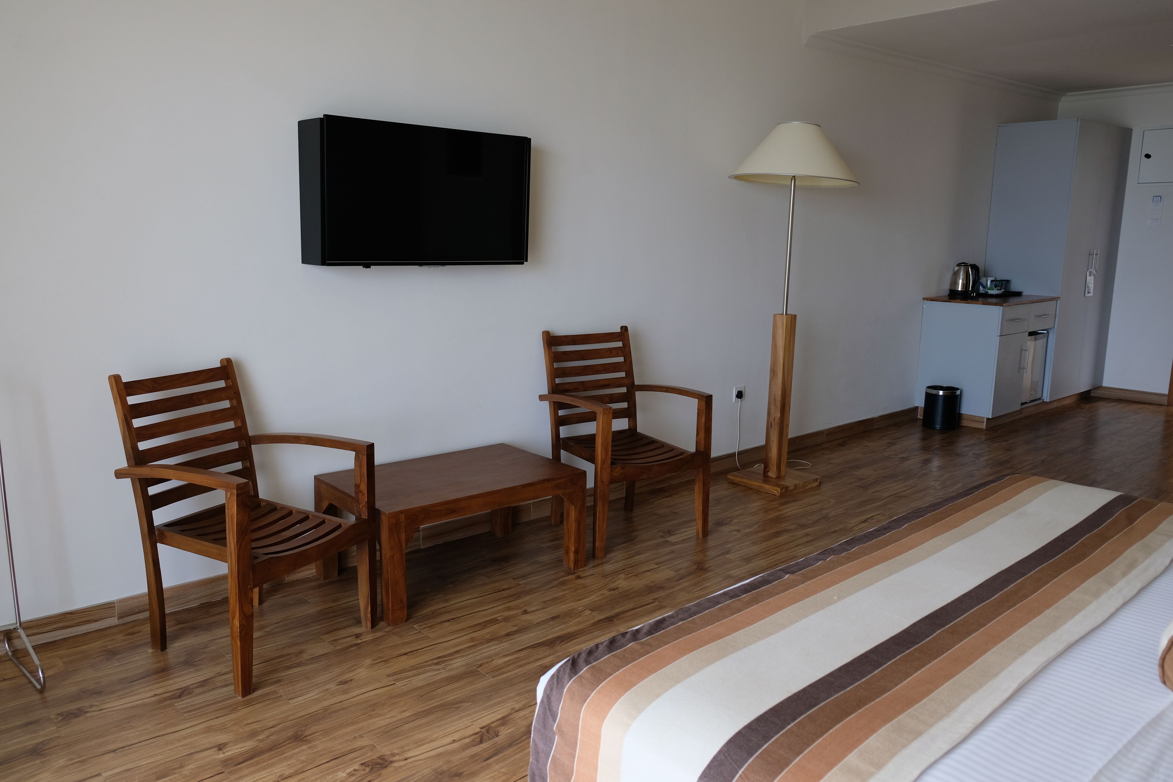 Una habitación con televisión montada en la pared, dos sillas de madera, una mesita, una lámpara de pie y una zona de kitchenette.