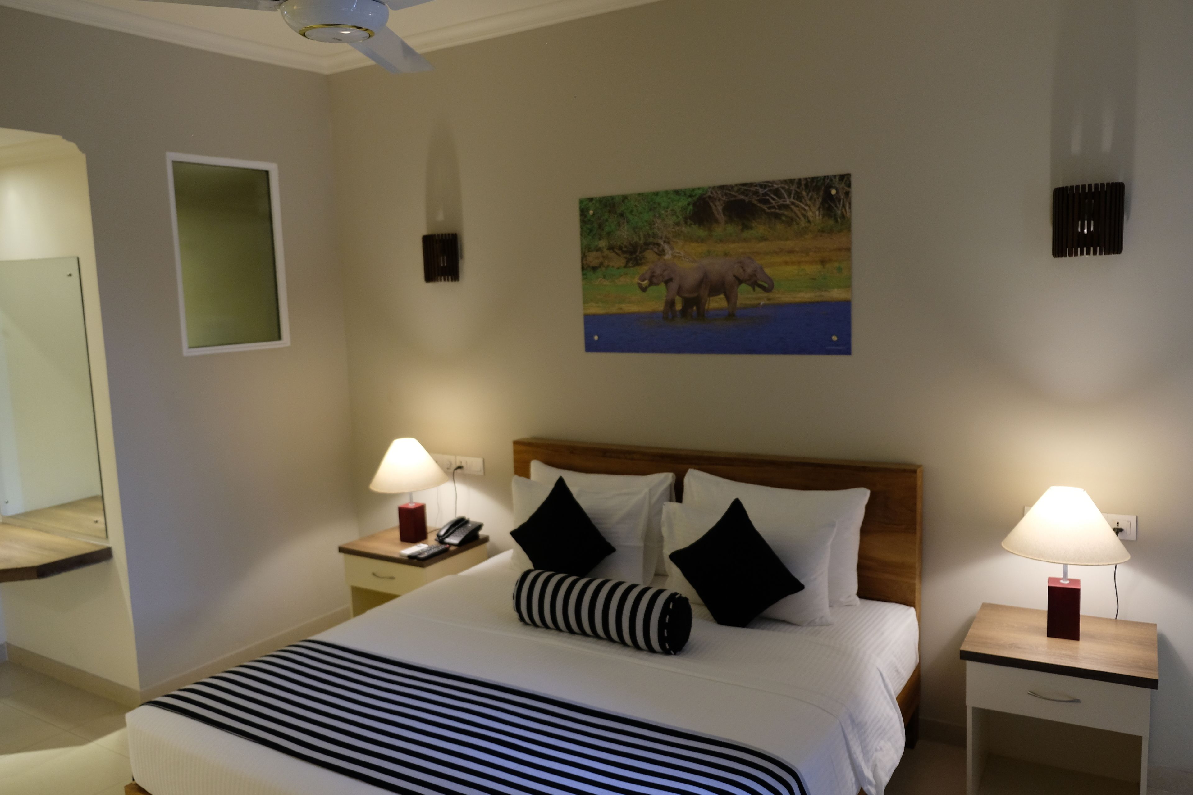 A hotel room with a large bed, two bedside tables with lamps, a wooden headboard, a painting of elephants, a mirror, and a small window.