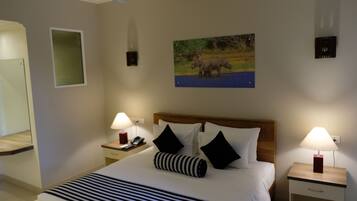 A hotel room with a large bed, two bedside tables with lamps, a wooden headboard, a painting of elephants, a mirror, and a small window.
