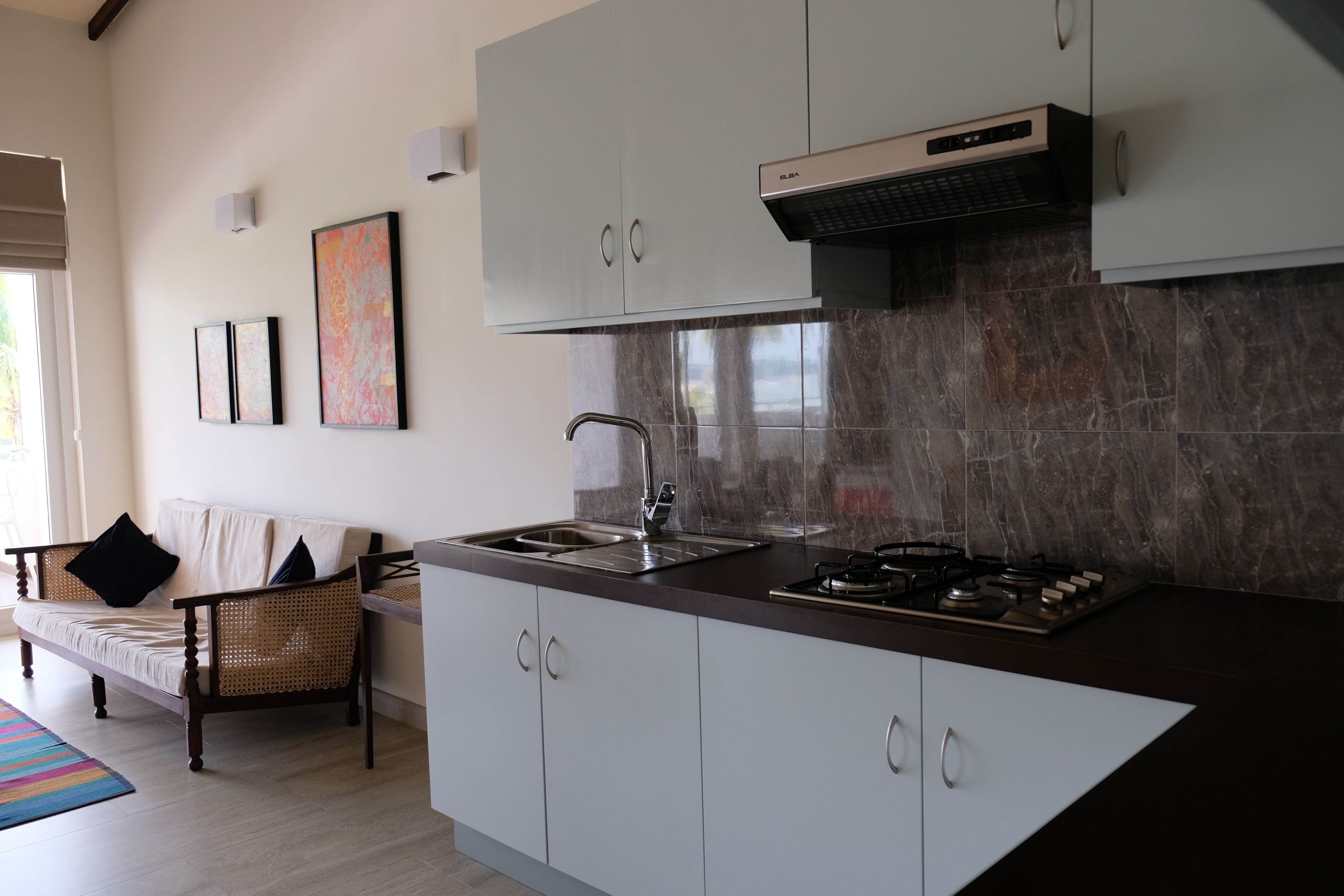 A modern kitchen with a built-in stove, a sink, and white cabinets. There is a dining area with a wicker chair and a colorful rug.