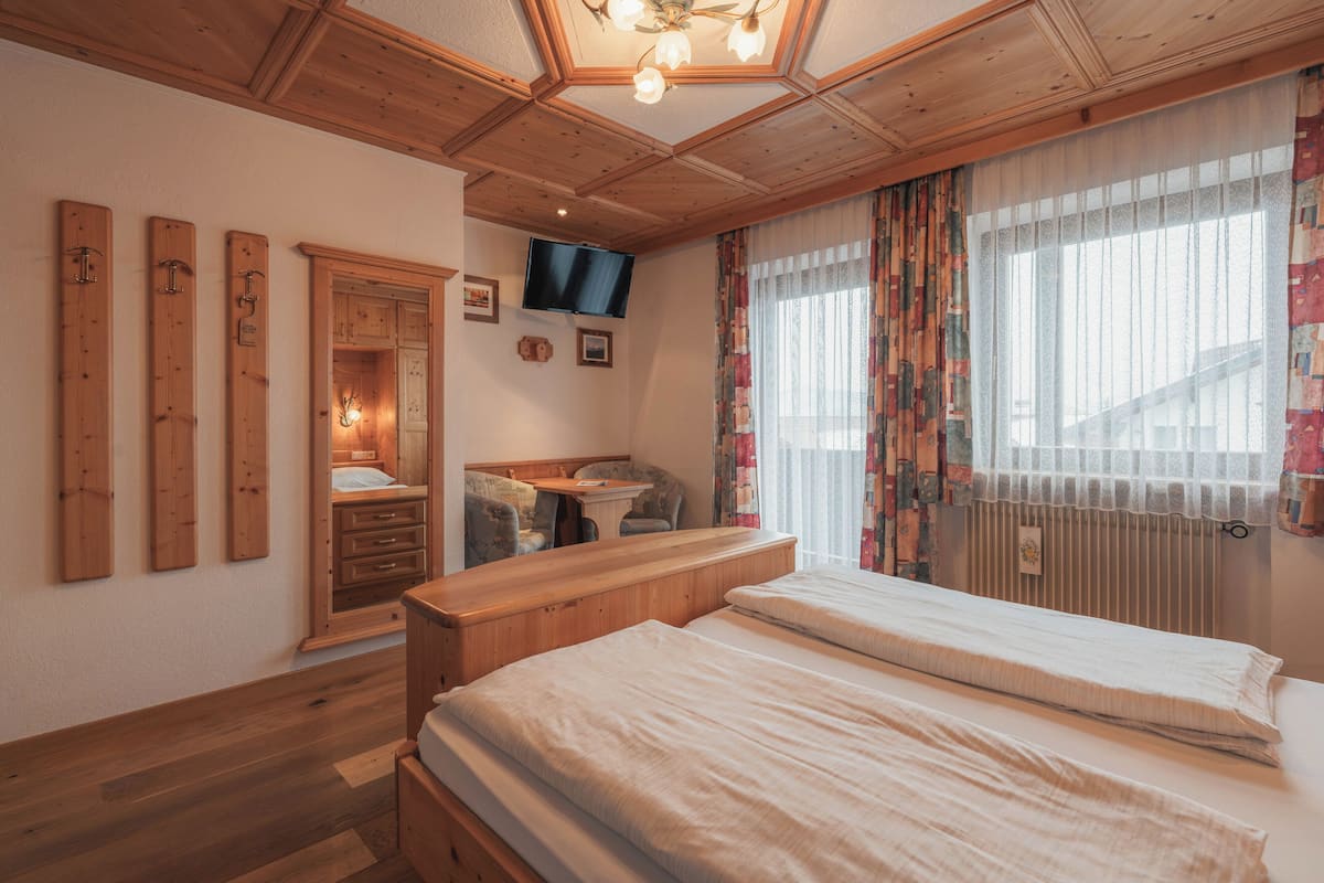 A bedroom with wooden flooring, a bed, a desk, a chair, a television, and a window with curtains.