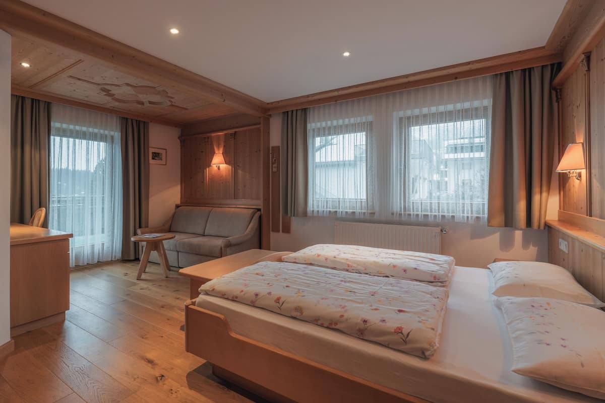 A room with a wooden ceiling, a bed with floral bedding, a sofa, a small table, and a window with curtains.