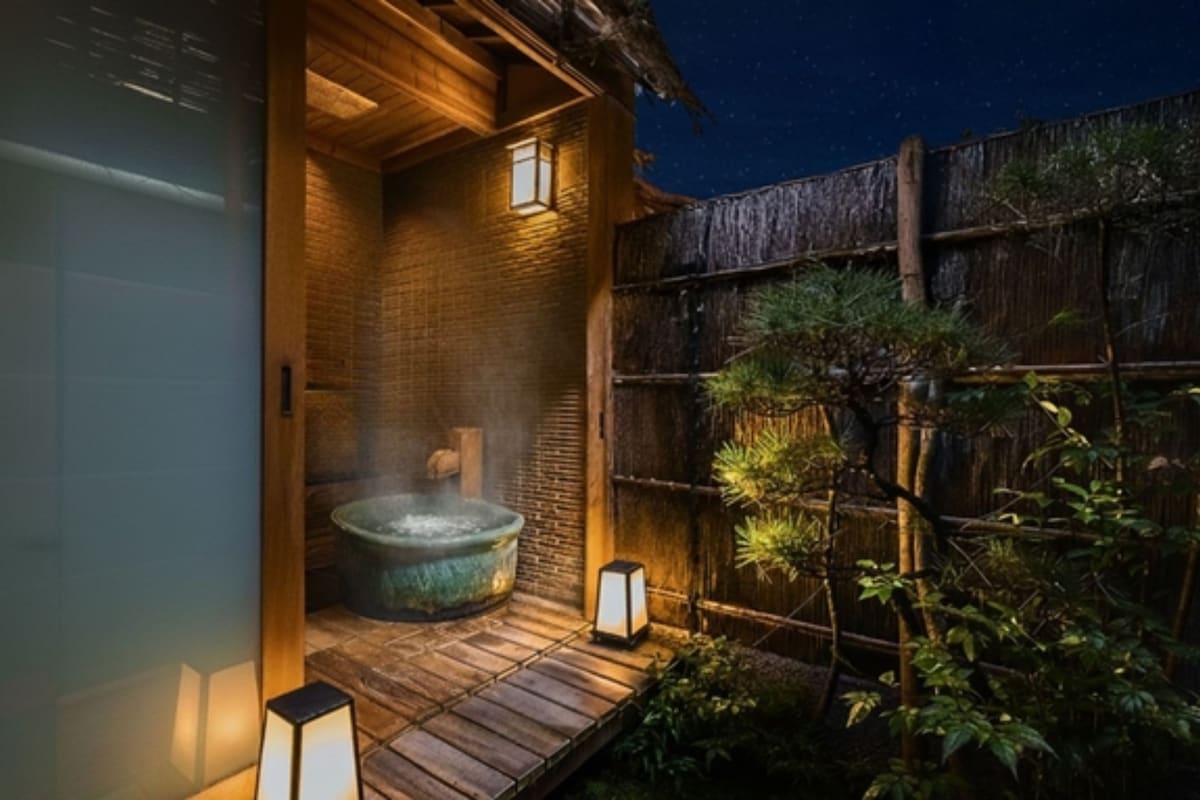 Un espace de source thermale en plein air de style japonais traditionnel, doté d’une terrasse en bois, d’un bain à remous en pierre et de deux lanternes.