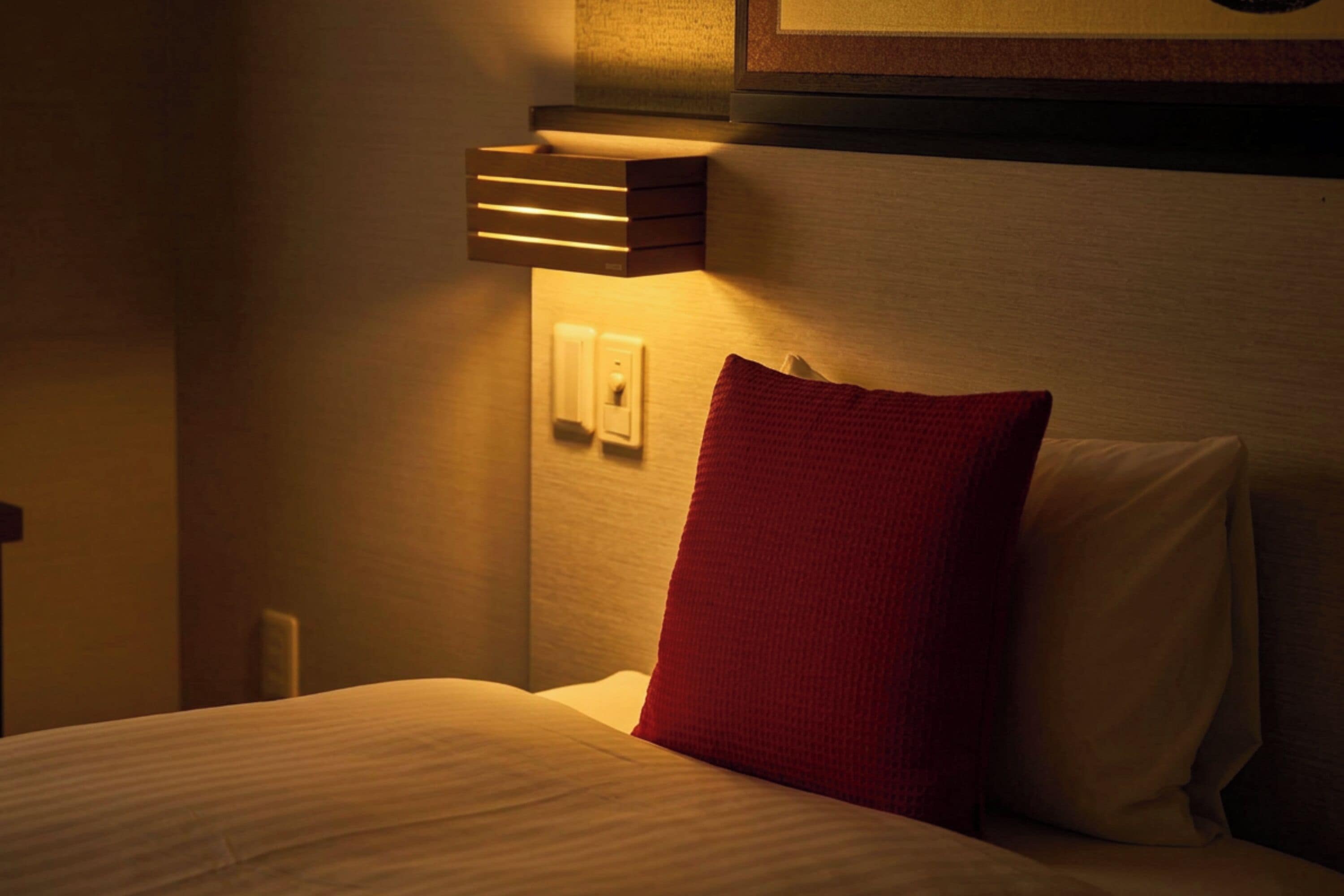 Una habitación de hotel con una cama, un almohadón rojo, una lámpara fijada a la pared y un enchufe.