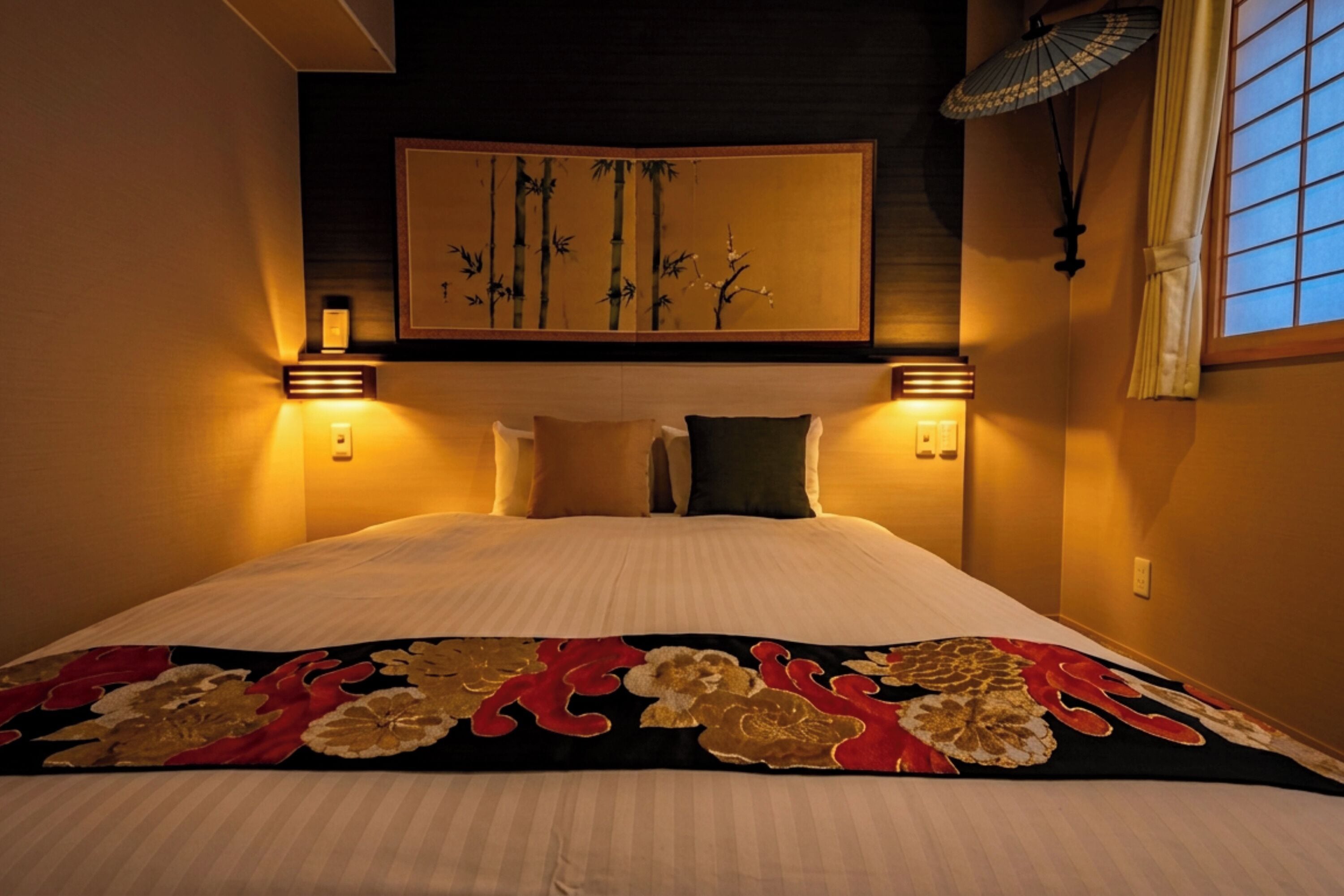 A hotel room with a bed, wall-mounted artwork, two wall sconces, a window with a view, and a decorative throw blanket.