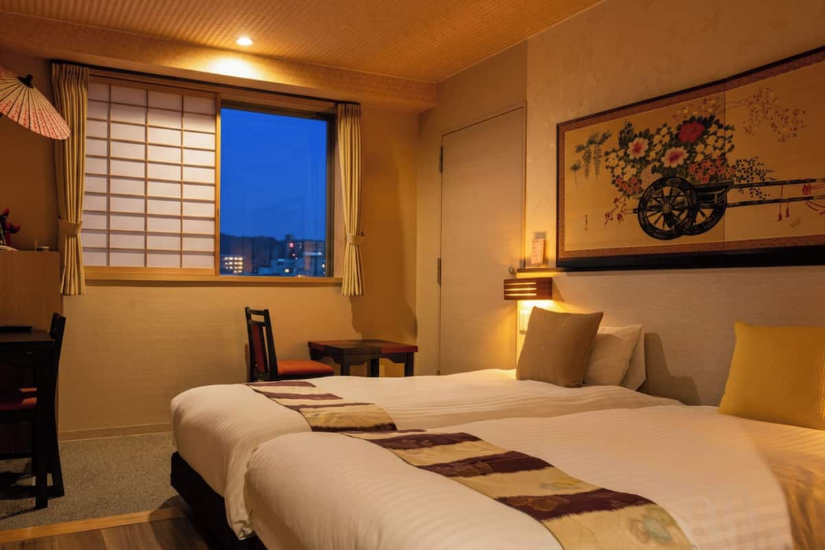 A hotel room with a large bed, a desk, a chair, a small table, a window with curtains, and a wall-mounted lamp.