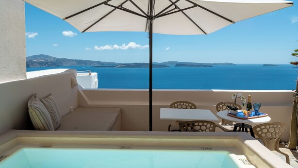 A hot tub with a view of the sea, a table with chairs, and an umbrella. - Passion Blue Villas (Santorini)