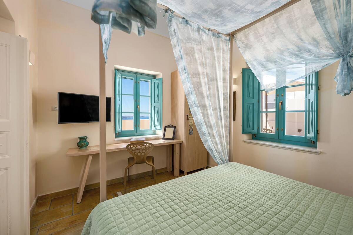 Un dormitorio con una cama, un escritorio con una silla, un televisor y dos ventanas con cortinas.