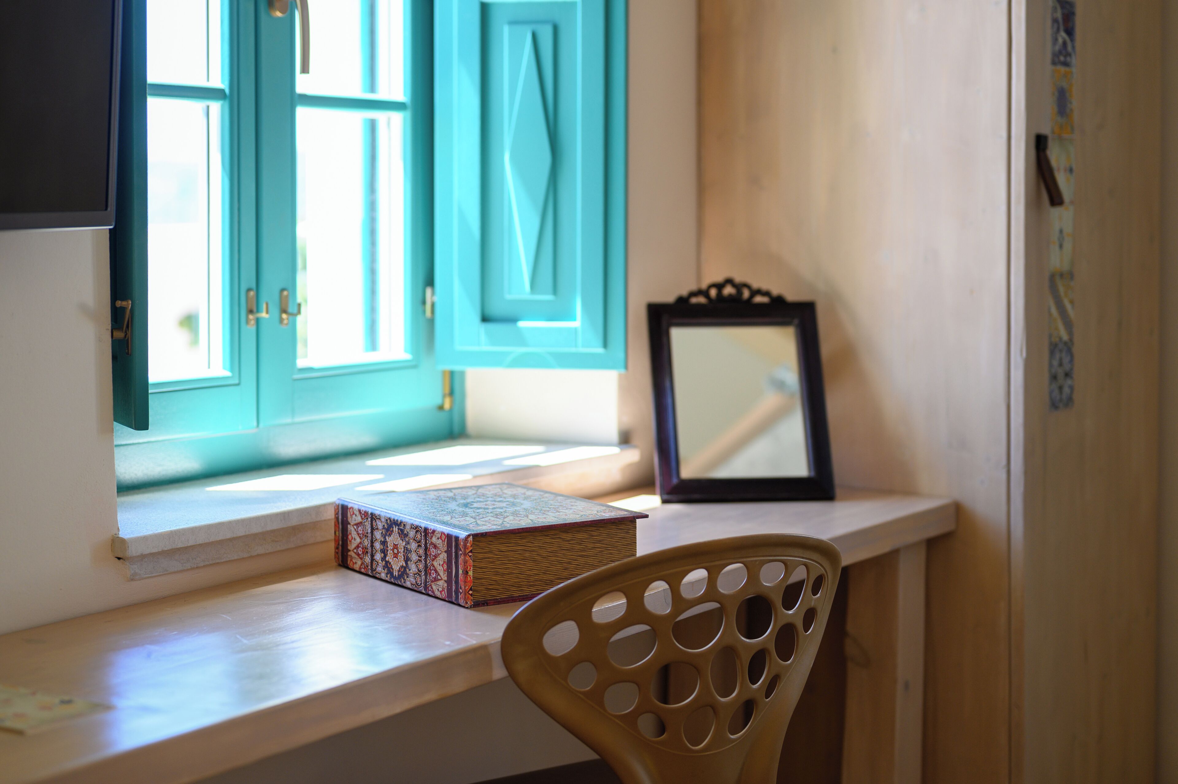 A desk with a book and a framed picture, a window with teal shutters, and a wooden chair.