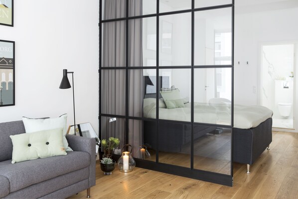 Une chambre moderne avec une cloison en verre, un canapé gris et une table de chevet avec une bougie.