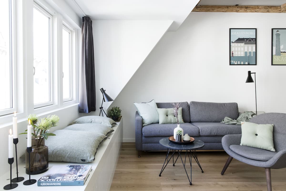 A modern living room with a grey sofa, a round coffee table, and a grey armchair. There is a window with curtains, a plant on the windowsill, and a book on the table.