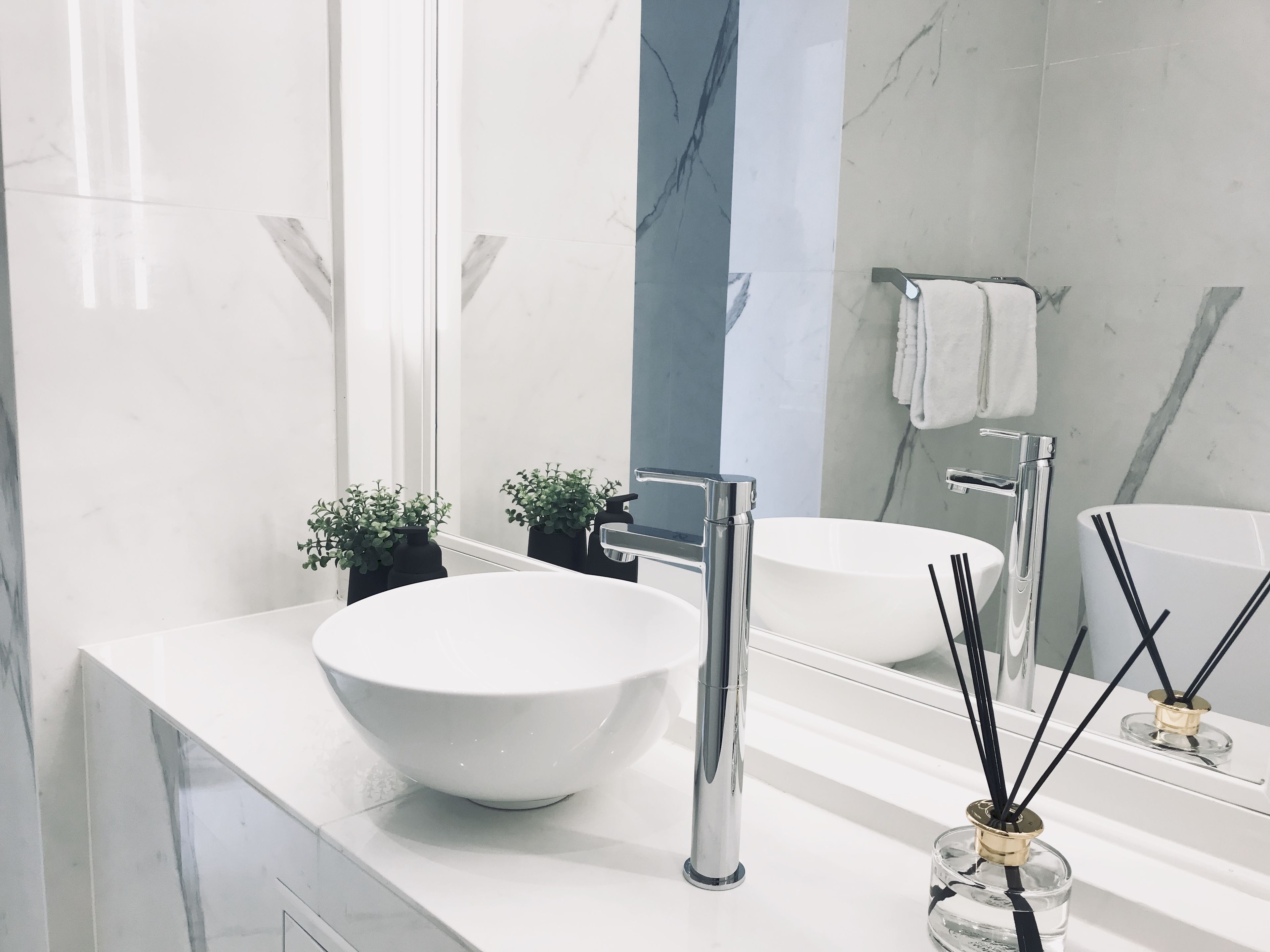 A modern bathroom with a white sink, chrome faucet, and a large mirror. There are two small potted plants on the counter.