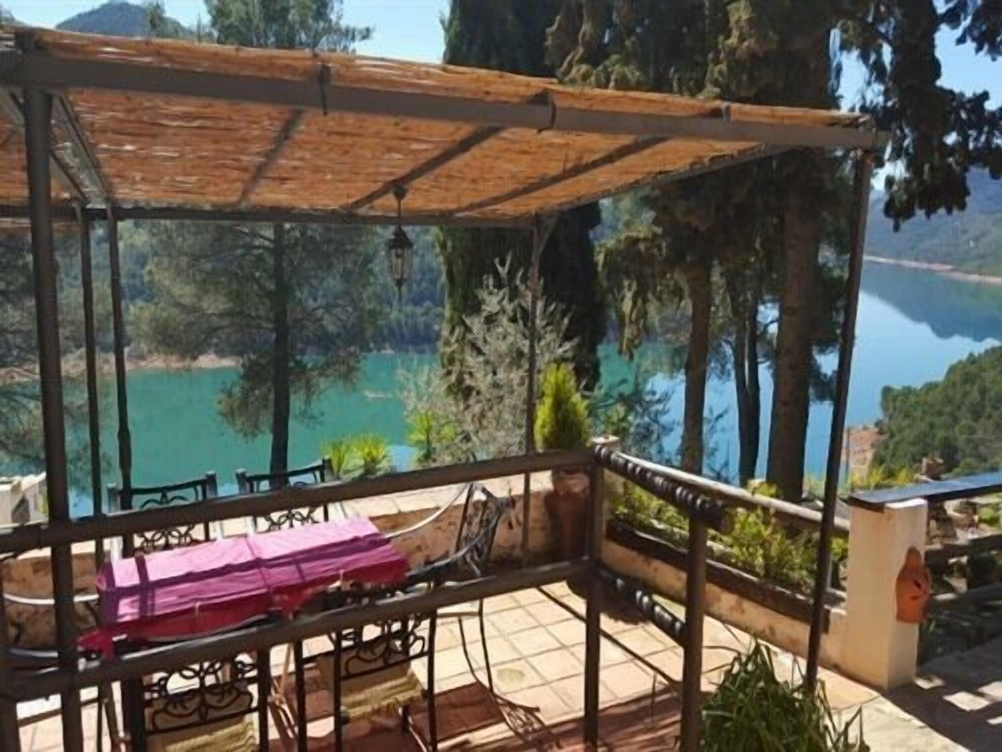 Un espacio al aire libre cubierto con mesa y sillas, con vistas a un lago y colinas boscosas.