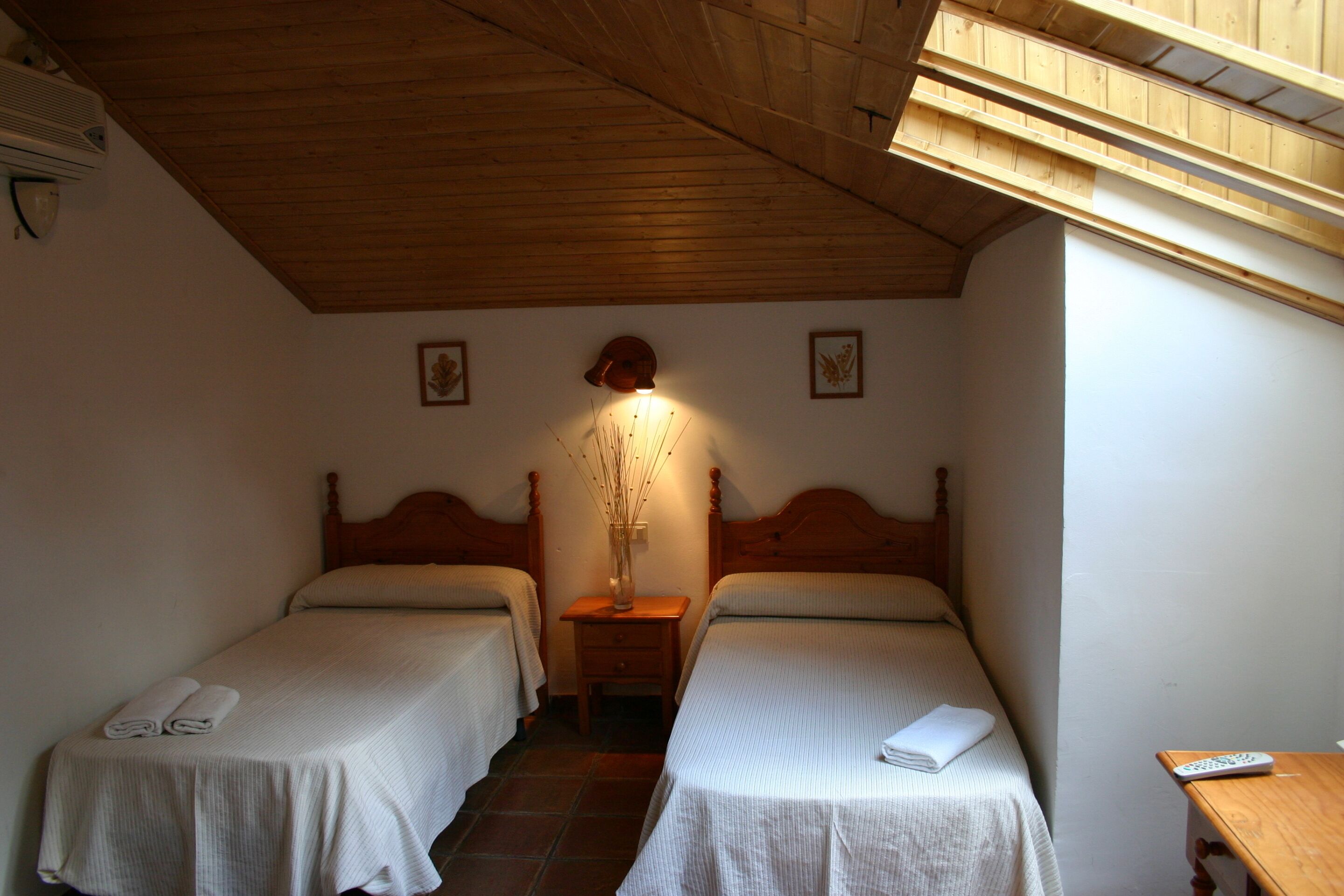 A small, cozy room with two single beds, a nightstand with a lamp, and a wooden ceiling.