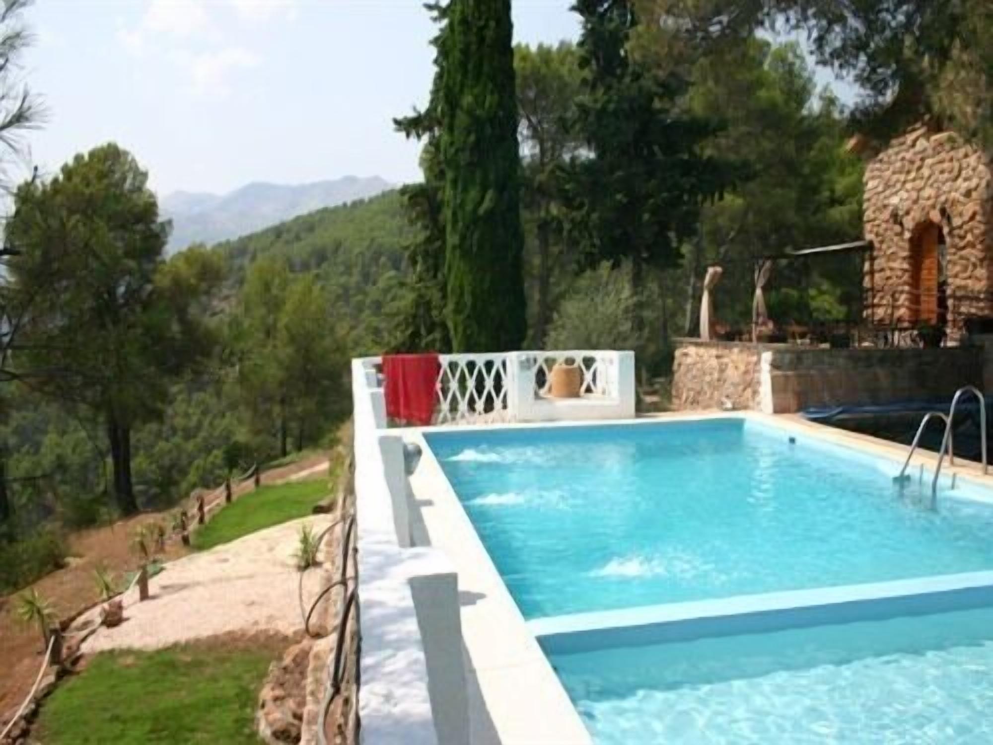 Una piscina rectangular con agua azul cristalina, rodeada por un solárium blanco y una cerca también blanca. Se observa una construcción de piedra con chimenea al fondo.