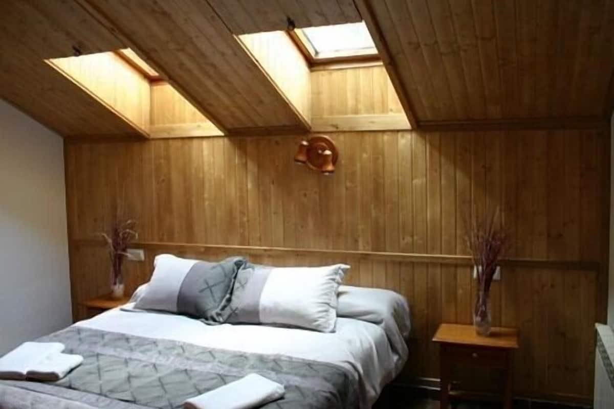 Ein Schlafzimmer mit Holzbalkendecke, einem Bett mit Kissen, einem Nachttisch und einer Vase mit Trockenpflanzen.
