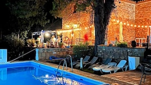Un área de piscina nocturna con muro de piedra, sillas reclinables y luces colgantes.