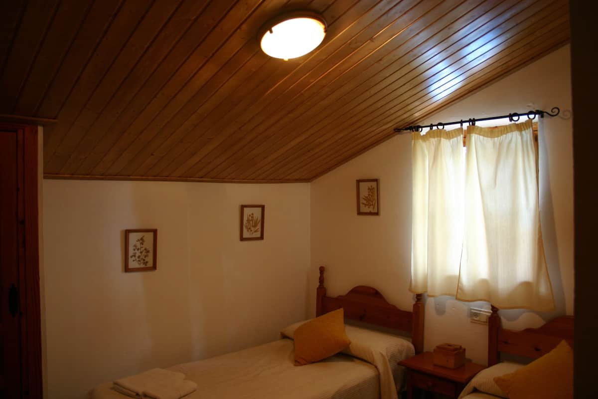 Ein Schlafzimmer mit zwei Einzelbetten, Holz-Kopfteilen, einer Decke mit Holzbalken und einem Fenster mit Vorhängen.