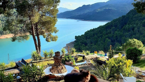 Una vista panorámica de un lago con montañas al fondo, rodeado de exuberante vegetación y una terraza de madera con mobiliario exterior.