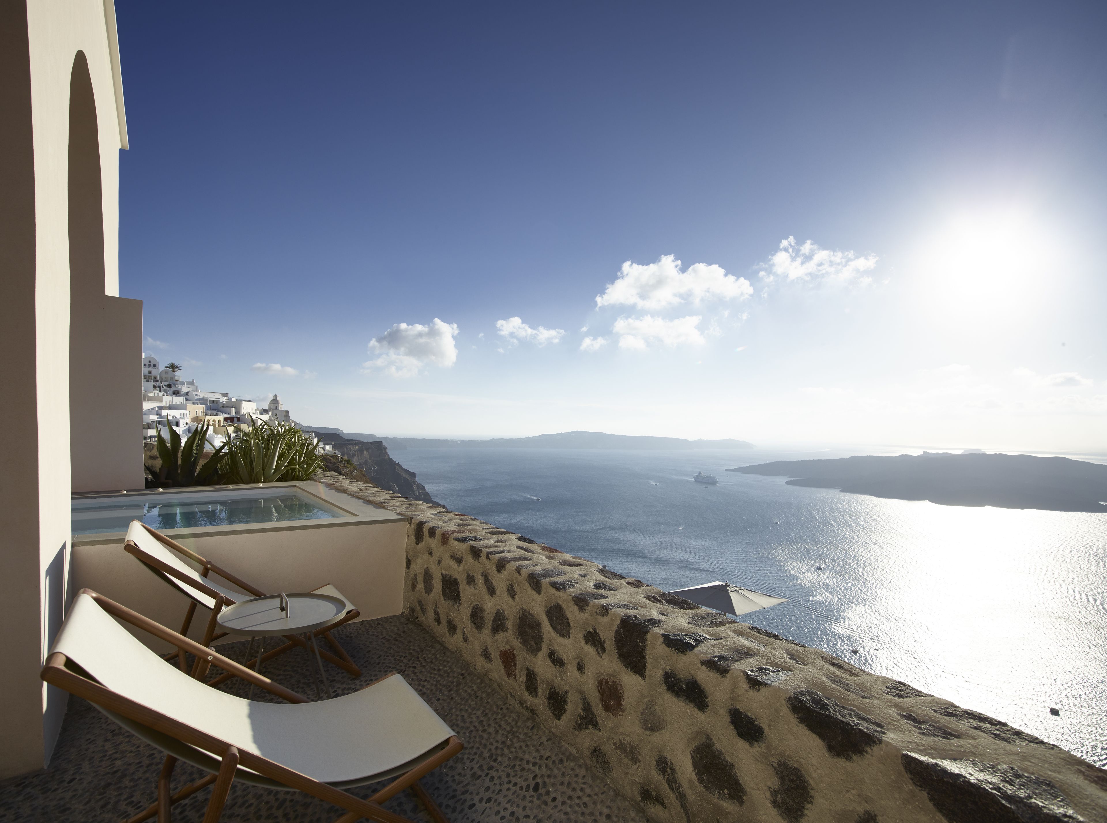 A balcony with a stone wall, a pool, and lounge chairs overlooking a coastal view.