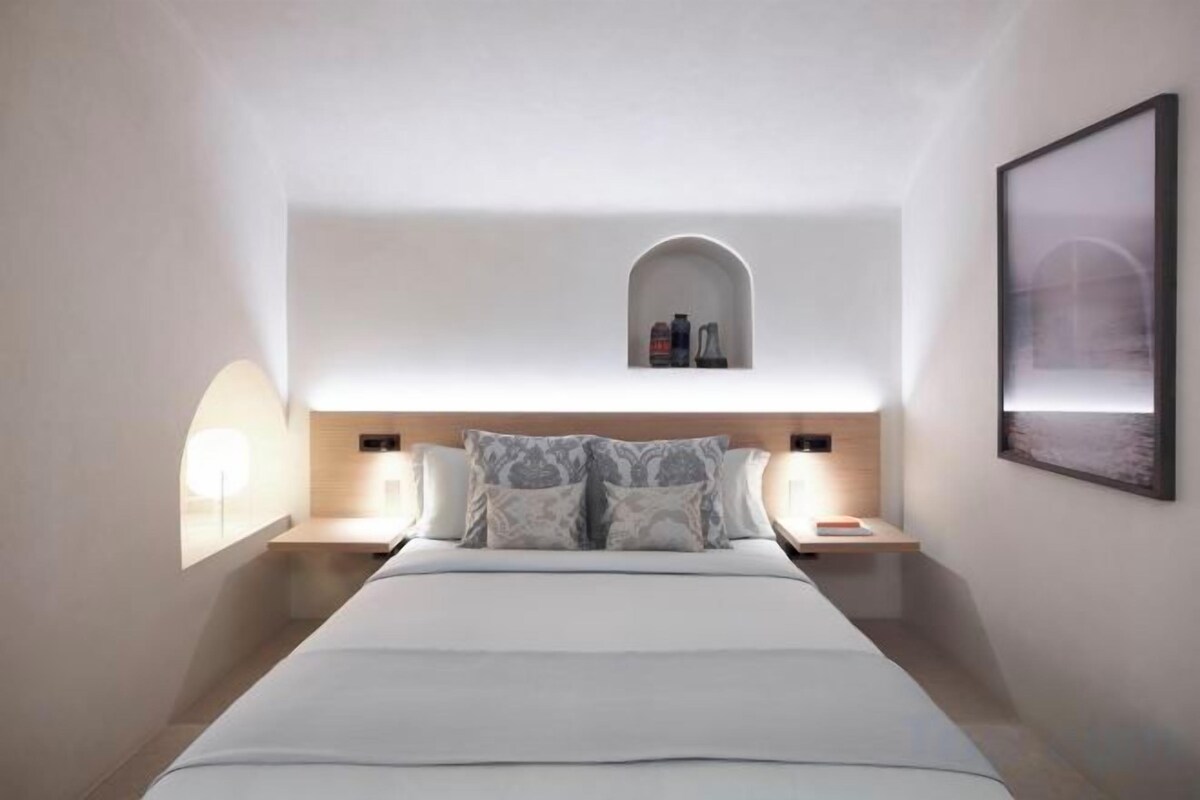 Habitación de hotel con una cama grande, lámparas de noche, un estante pequeño y un cuadro en la pared.