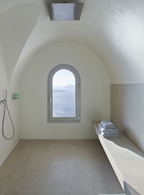 A small, private bathroom with a shower, a window with a view of the sea, and a shelf with towels.