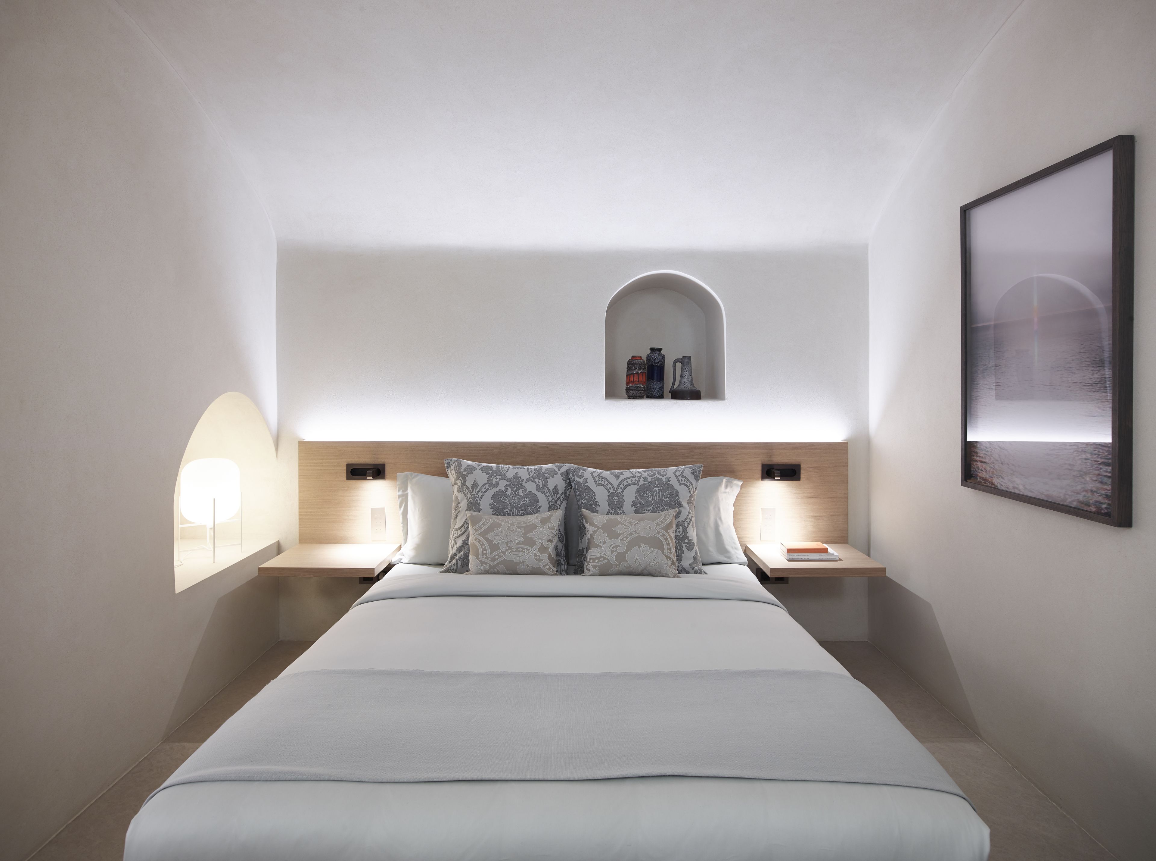 A modern hotel room with a large bed, two bedside tables, a small shelf with decorative items, and a framed picture on the wall.