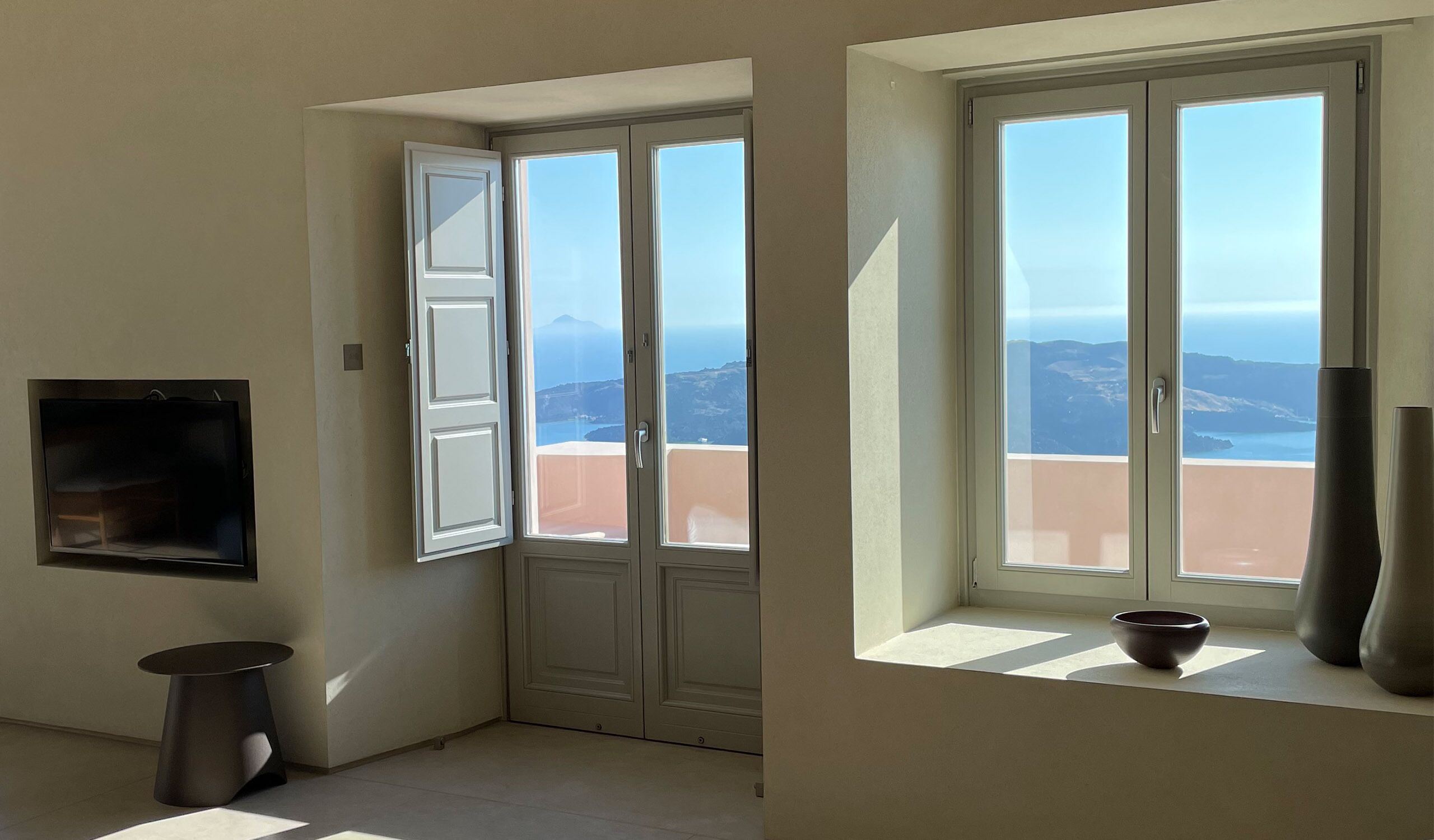 A room with a large sliding glass door leading to a balcony with a view of the sea and mountains. There is a small table with a bowl on it and a wall-mounted TV.