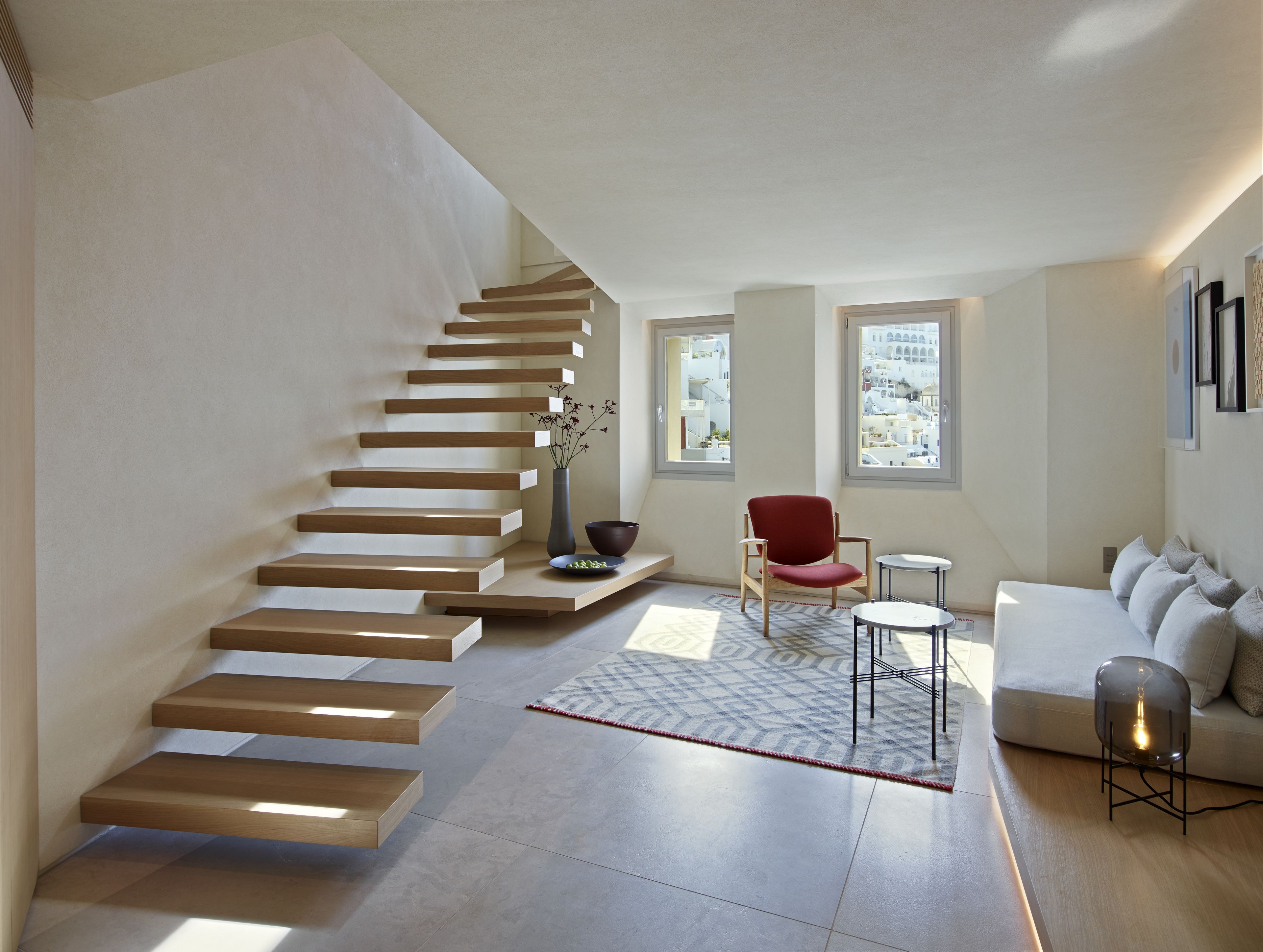 A modern living room with a wooden staircase, a red chair, a white sofa, and a view of a cityscape through large windows.