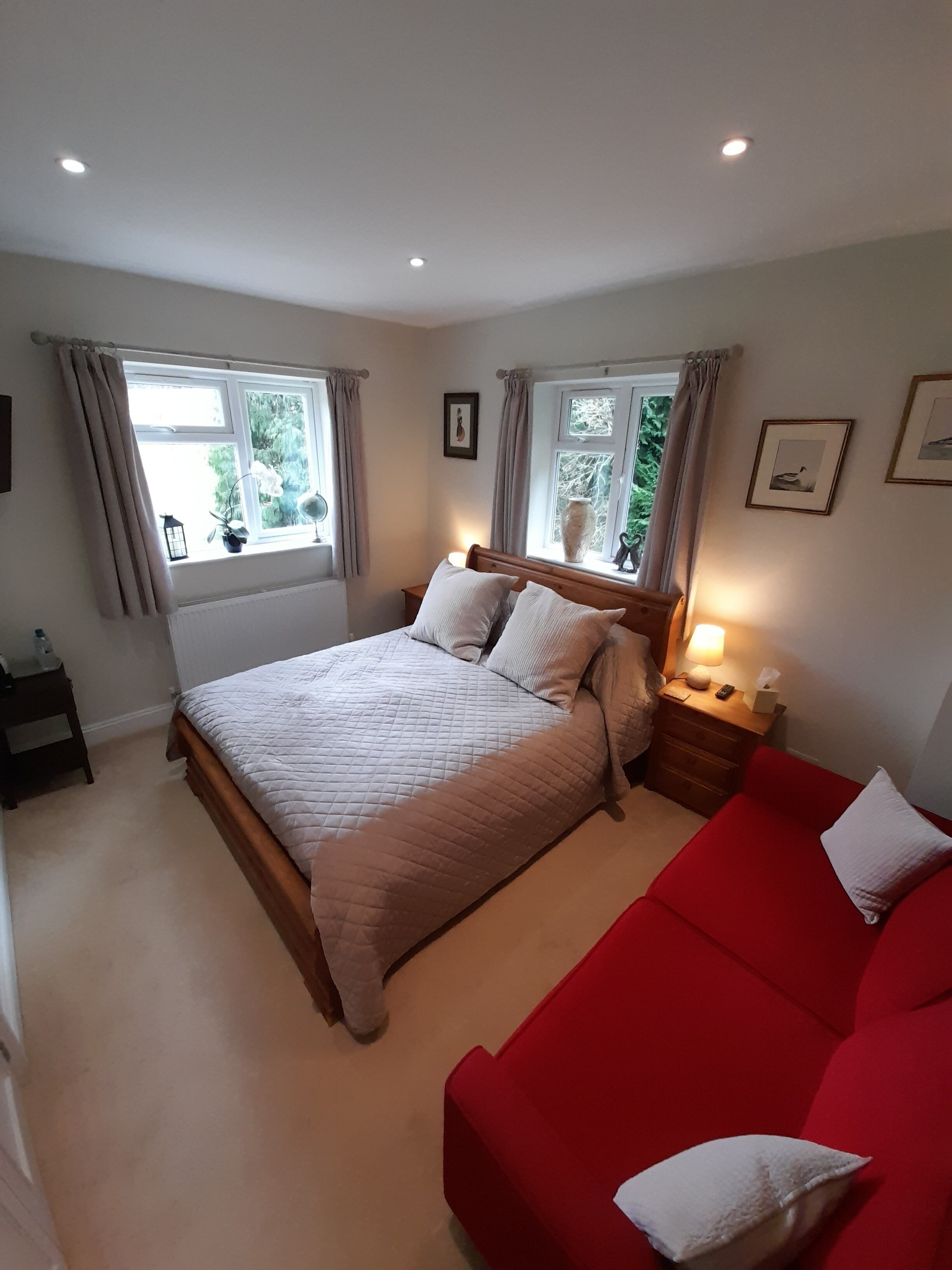 A bedroom with a bed, a red sofa, a nightstand, and two windows with curtains.