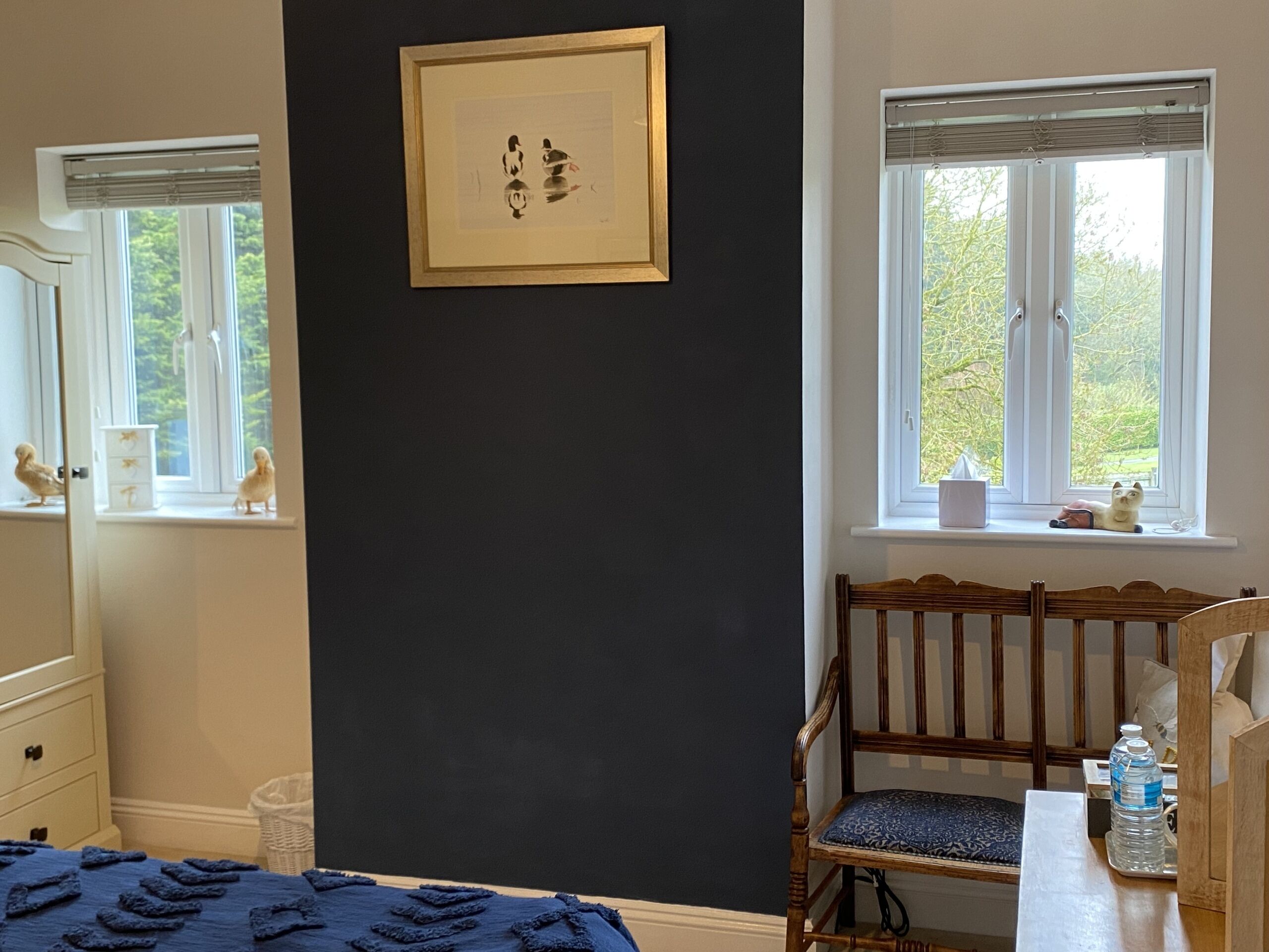 A room with a dark wall, a framed artwork, a wooden chair, a bed, a window with a view of trees, and a small shelf with items.
