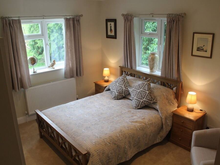 Un dormitorio con cama, dos lámparas de noche, una mesita de noche, una silla y dos ventanas con cortinas.