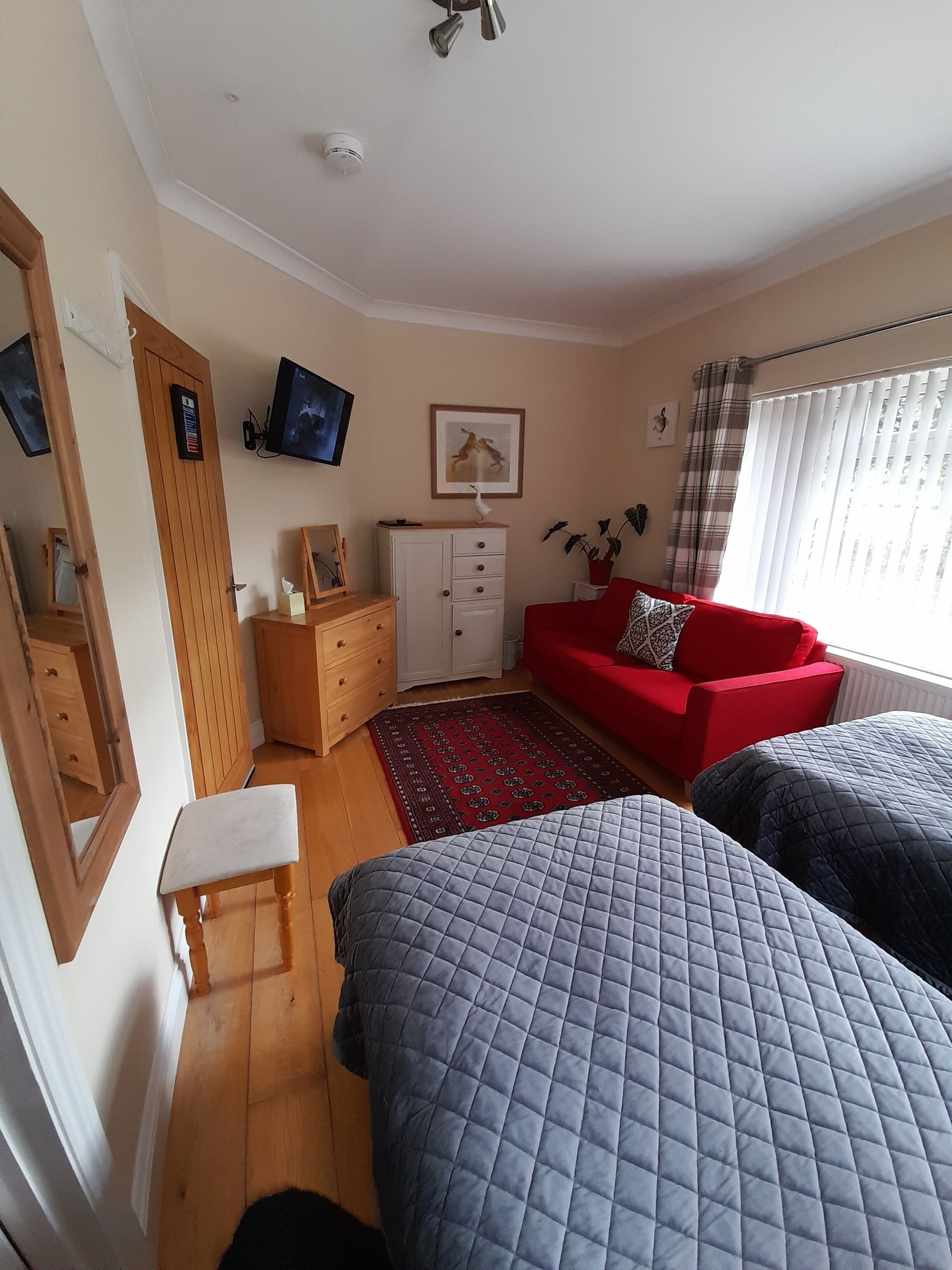 A room with two beds, a red sofa, a wooden dresser, a television, and a window with curtains.