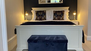 A bedroom with a large bed, a bedside table, a nightstand, a dark blue bench, and a mirror on the wall. - Sunny Nest (Cheltenham)