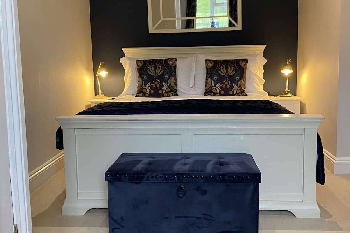 A bedroom with a large bed, a bedside table, a nightstand, a dark blue bench, and a mirror on the wall.