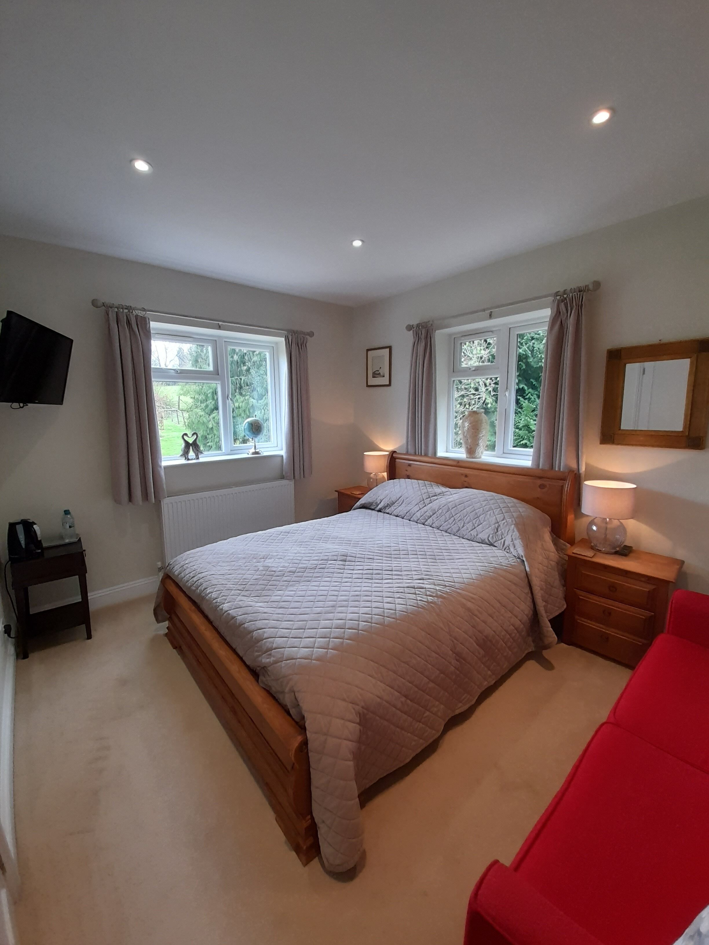 A bedroom with a bed, two bedside tables, a red chair, a TV mounted on the wall, and windows with curtains.