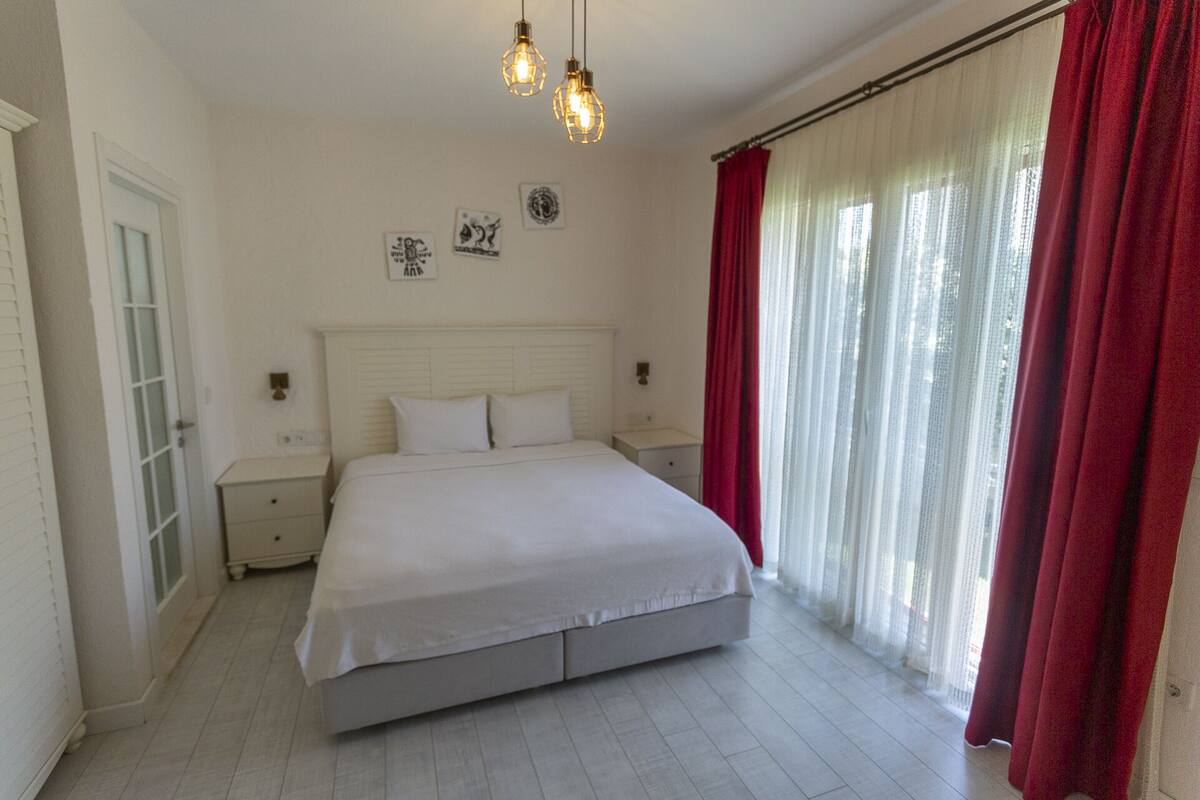 A bedroom with a large bed, white curtains, and a window with red drapes.