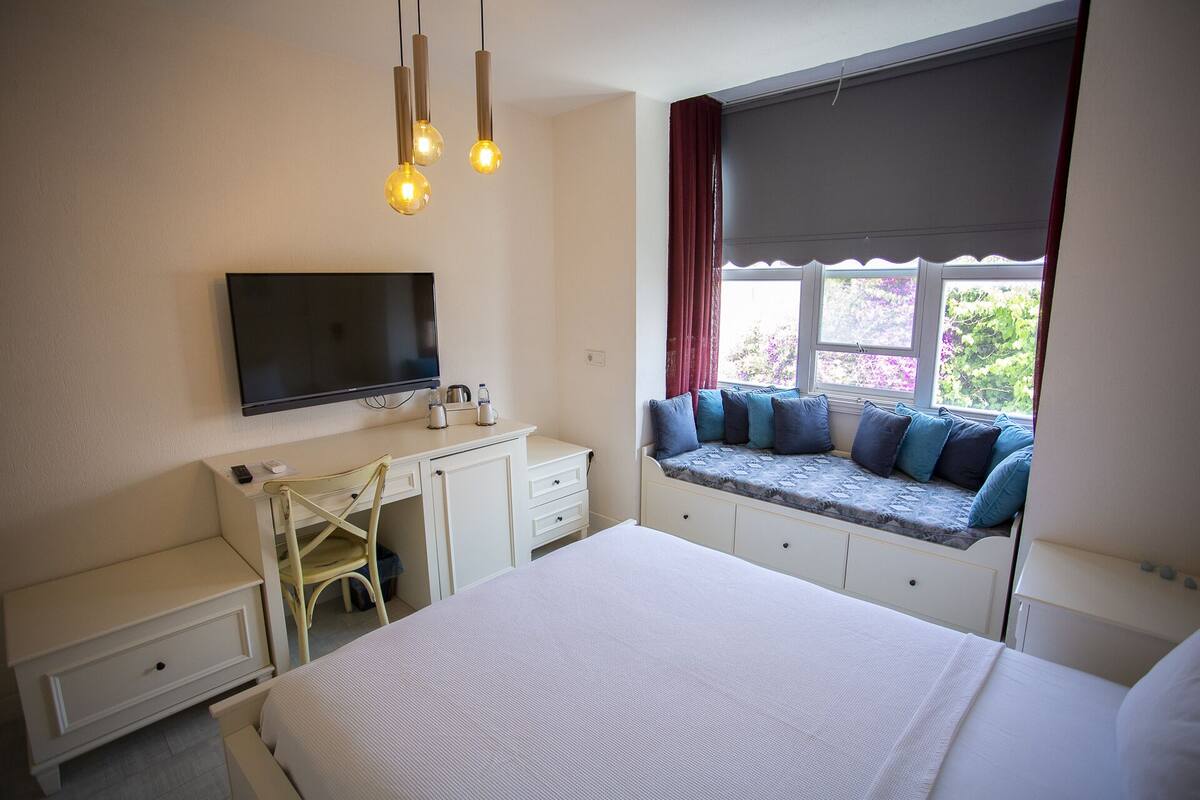 A bedroom with a bed, a desk with a TV, a window seat with cushions, and a chair.