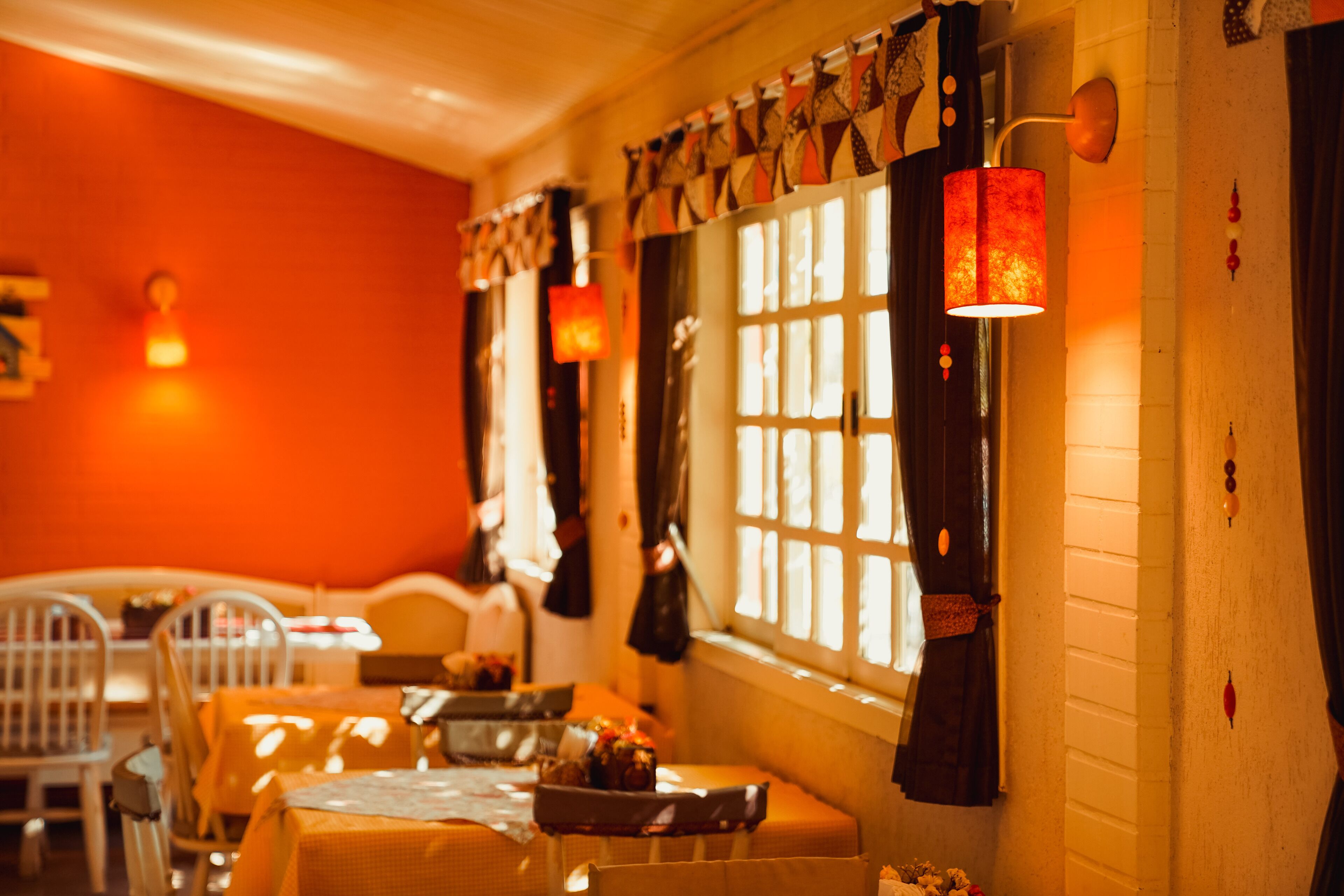 A cozy restaurant with a warm interior, featuring a dining area with tables and chairs, a window with curtains, and wall-mounted lighting.