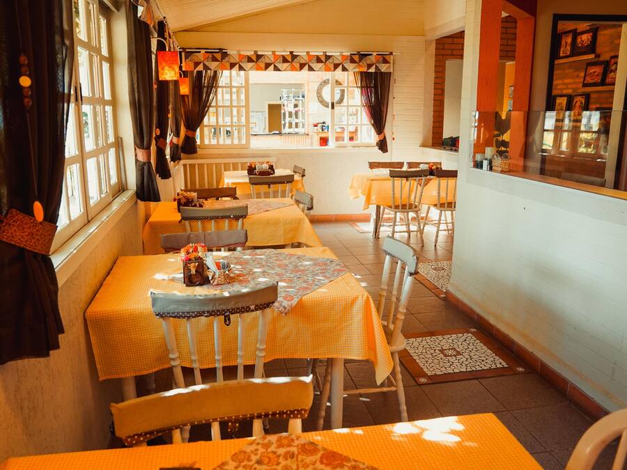 Un restaurante acogedor con varias mesas y sillas, vista a la calle a través de amplios ventanales, y una pared de ladrillo con cuadros enmarcados.