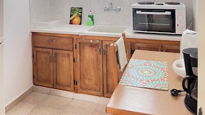 Uma cozinha pequena com armários de madeira, bancada branca, micro-ondas, máquina de café e uma janela com cortinas leves.