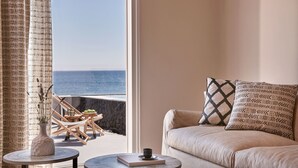 A room with a view of the sea, a sofa with patterned pillows, a small table with a book and a cup, and a wicker chair by the window. - Incognito Villa (Santorini)
